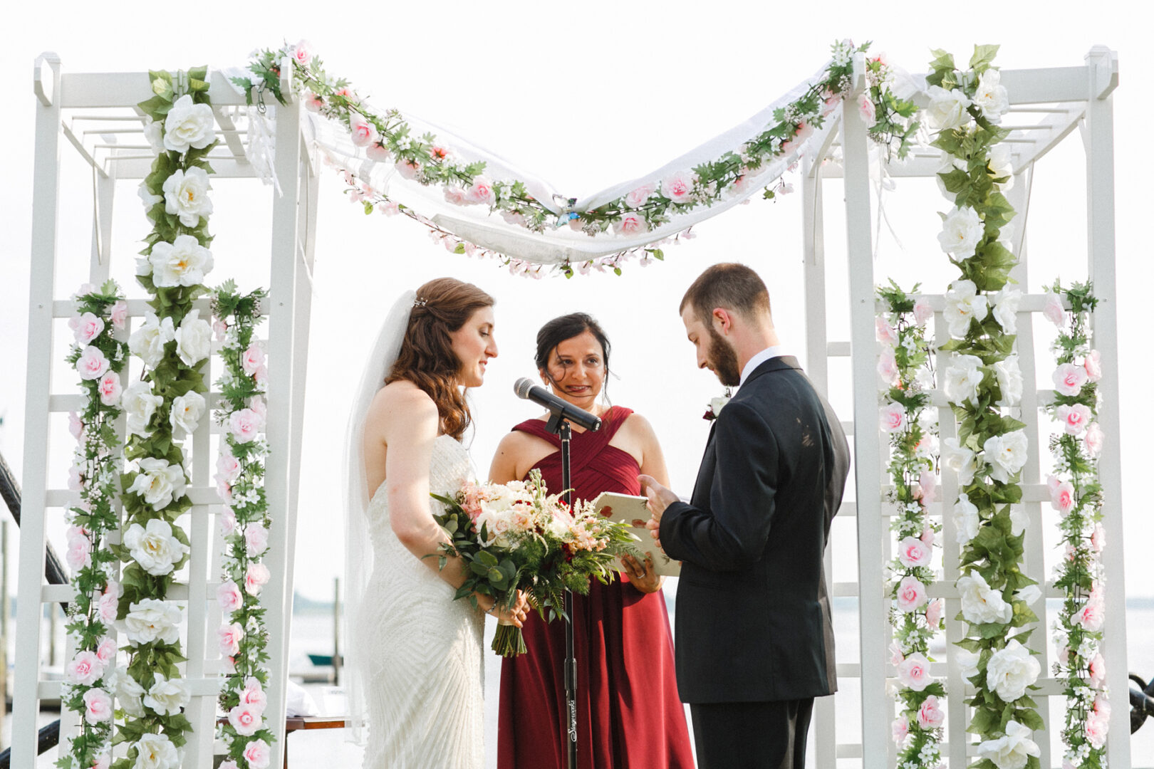 A bride and groom stand under a floral arch during an outdoor wedding ceremony at the Corinthian Yacht Club of Philadelphia, with an officiant in a red dress reading from a book.
