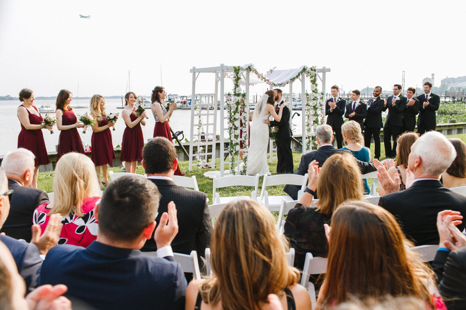 A bride and groom kiss under a floral arch at an outdoor wedding ceremony at the Corinthian Yacht Club of Philadelphia. Bridesmaids in red dresses and groomsmen in suits stand on either side as guests seated nearby clap with joy.