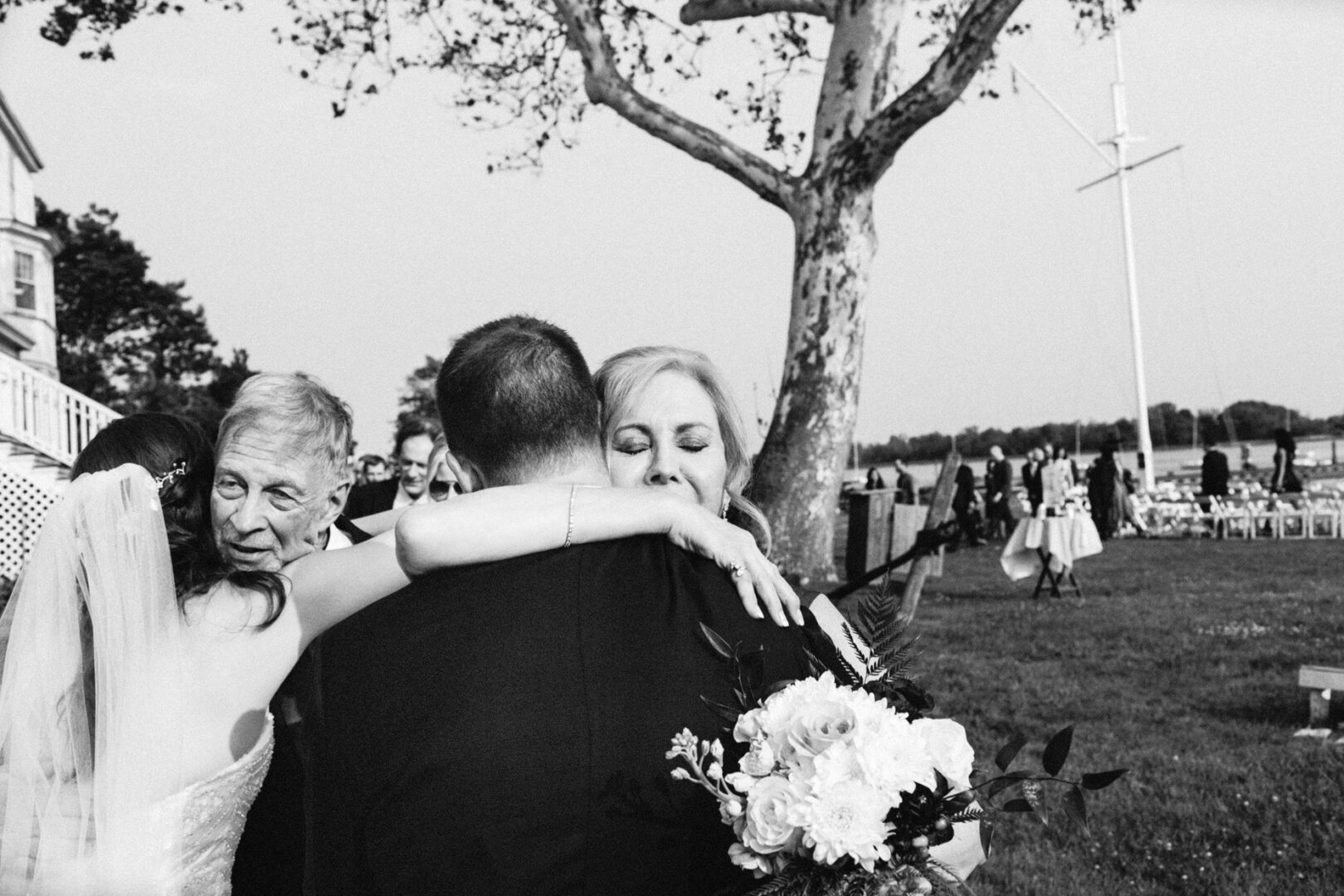 A bride embraces a man with a bouquet at the Corinthian Yacht Club of Philadelphia wedding, while an elderly man looks on. Guests and chairs are scattered in the background near a tree.