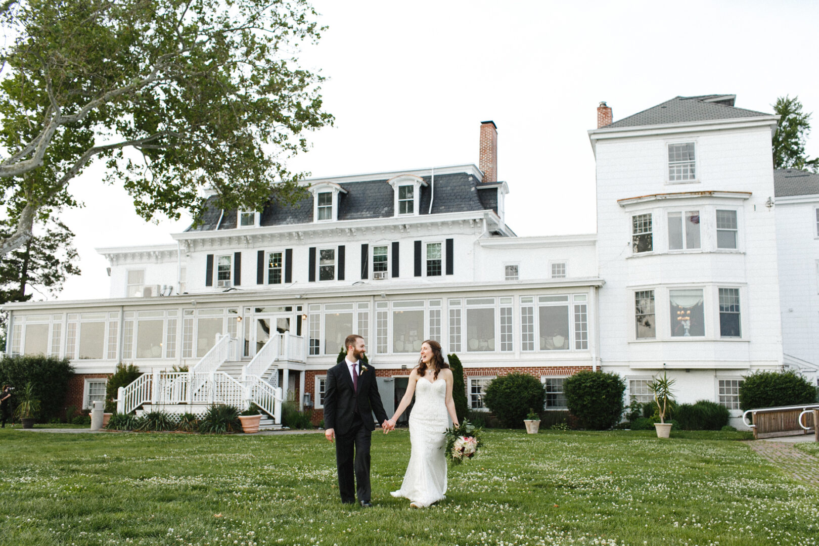 A couple in wedding attire holds hands, strolling across a lush lawn in front of the grand Corinthian Yacht Club of Philadelphia. The bride, with a bouquet in hand, beams as sunlight dances off the house’s many windows.
