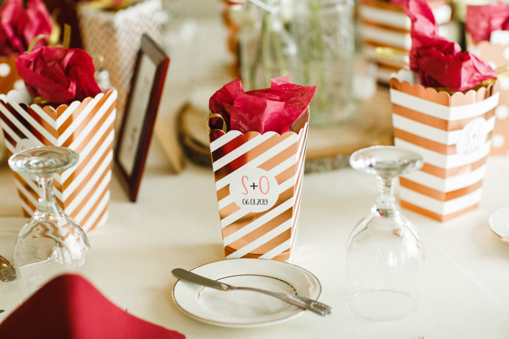 Table setting at a Corinthian Yacht Club of Philadelphia wedding features striped popcorn boxes filled with red tissue paper, surrounded by upside-down wine glasses and a small plate with cutlery.