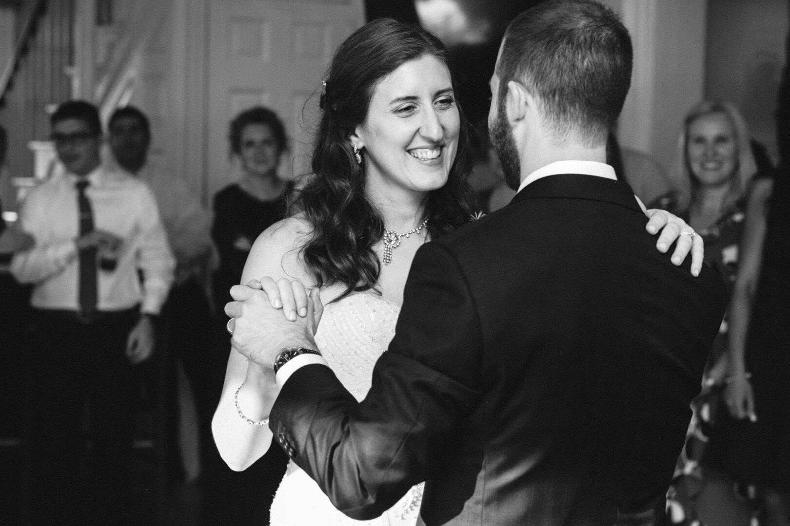 A bride and groom share a dance at their Corinthian Yacht Club of Philadelphia wedding reception, surrounded by smiling guests. The image is in black and white.