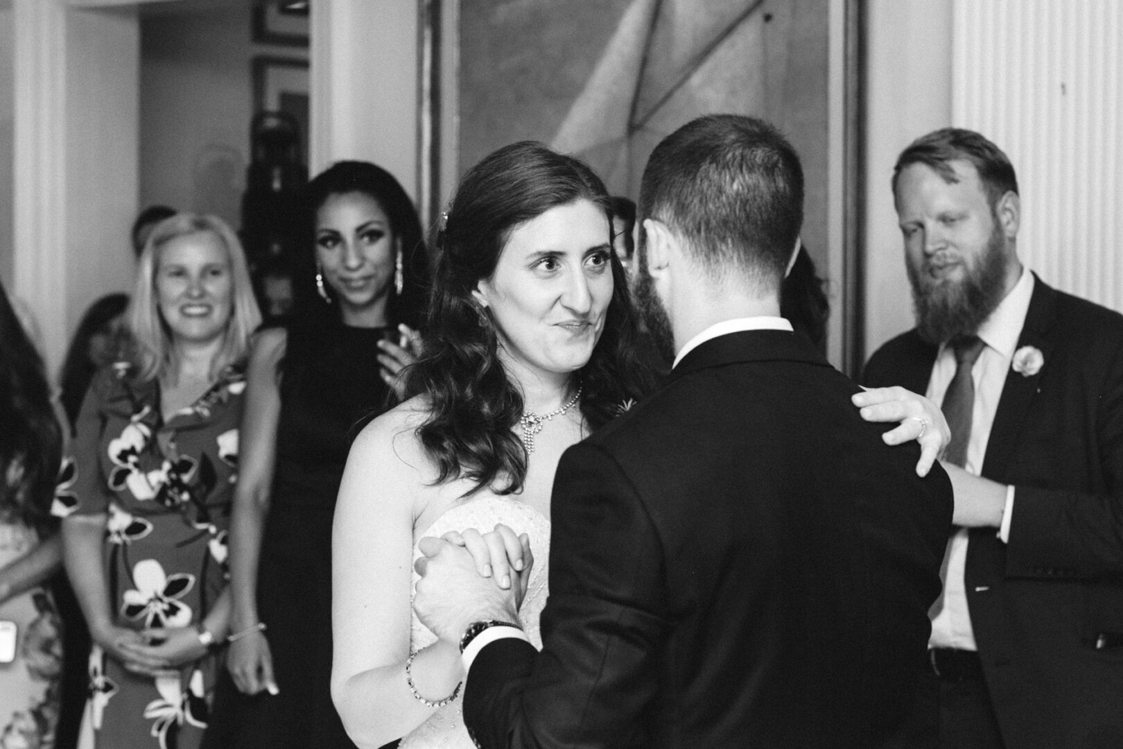 Amidst the elegance of a Corinthian Yacht Club of Philadelphia wedding, a couple dances closely, surrounded by smiling guests. The indoor setting is adorned with framed art, adding to the sophisticated ambiance of this joyous occasion.