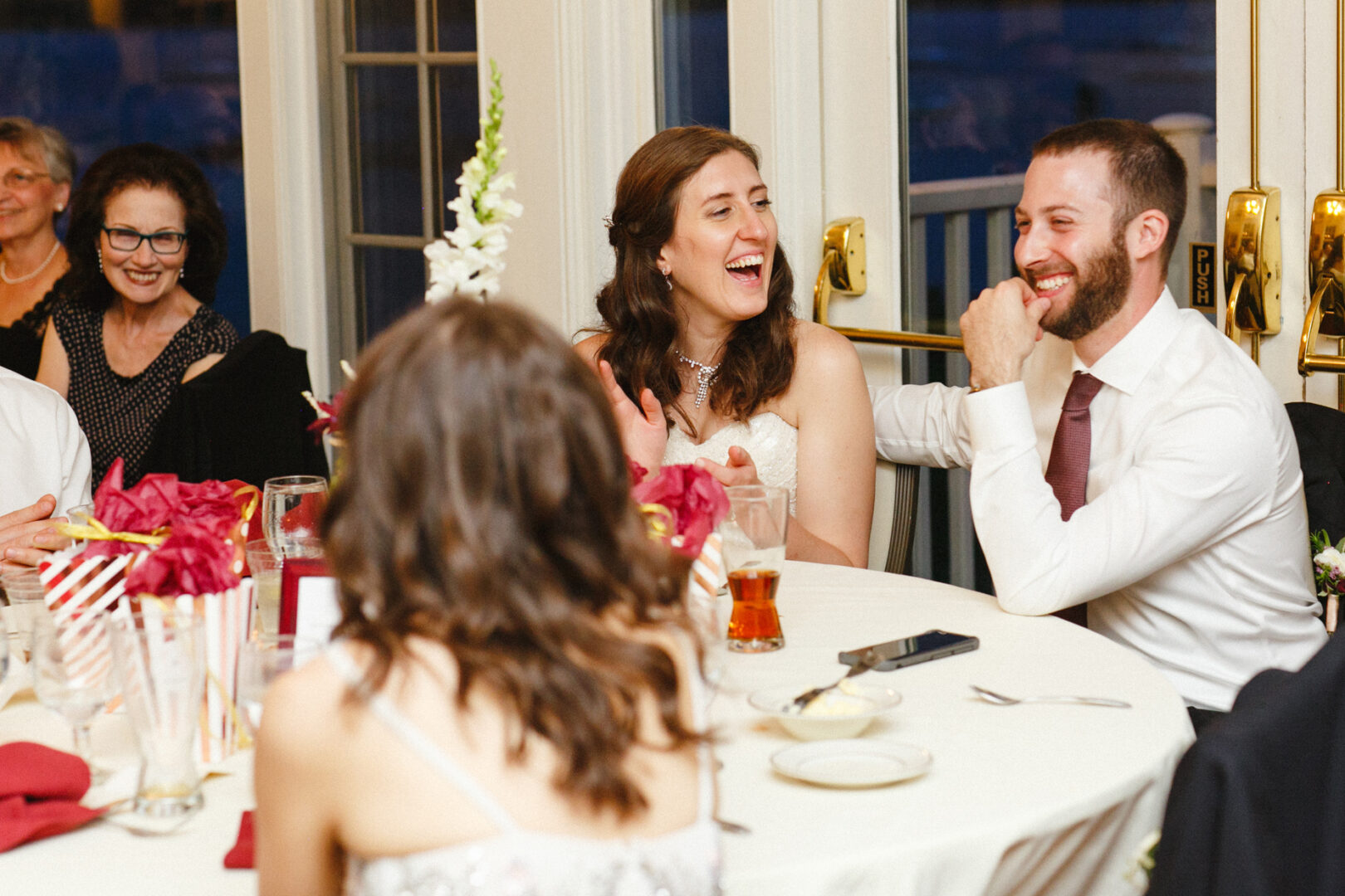 At the Corinthian Yacht Club of Philadelphia wedding, people sat around a table at an indoor event, laughing and chatting. A woman and a man were at the center, surrounded by elegant decorations and drinks on the table.