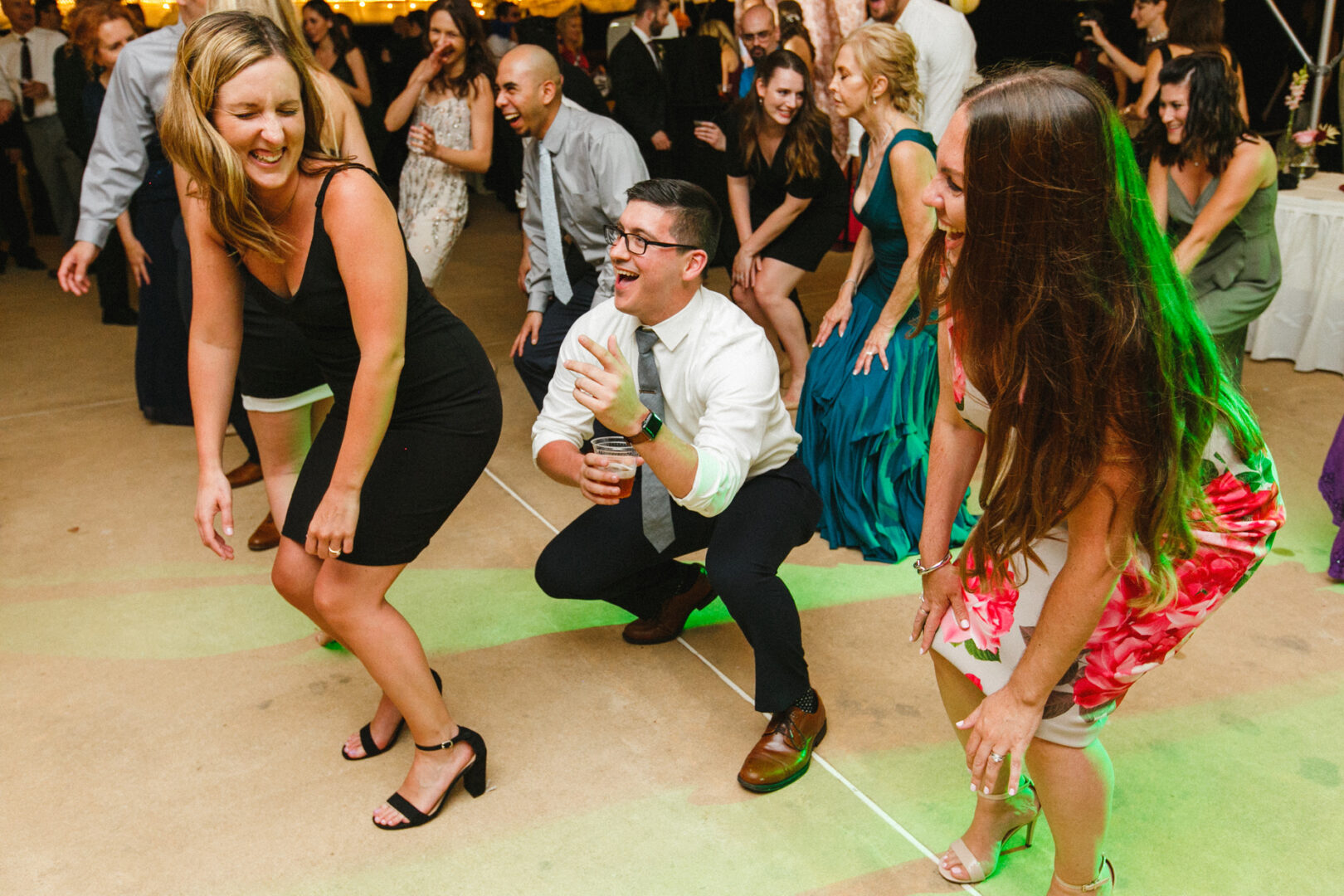 At a lively event reminiscent of a Corinthian Yacht Club of Philadelphia wedding, people are dancing energetically indoors, with one man playfully squatting as others laugh heartily.