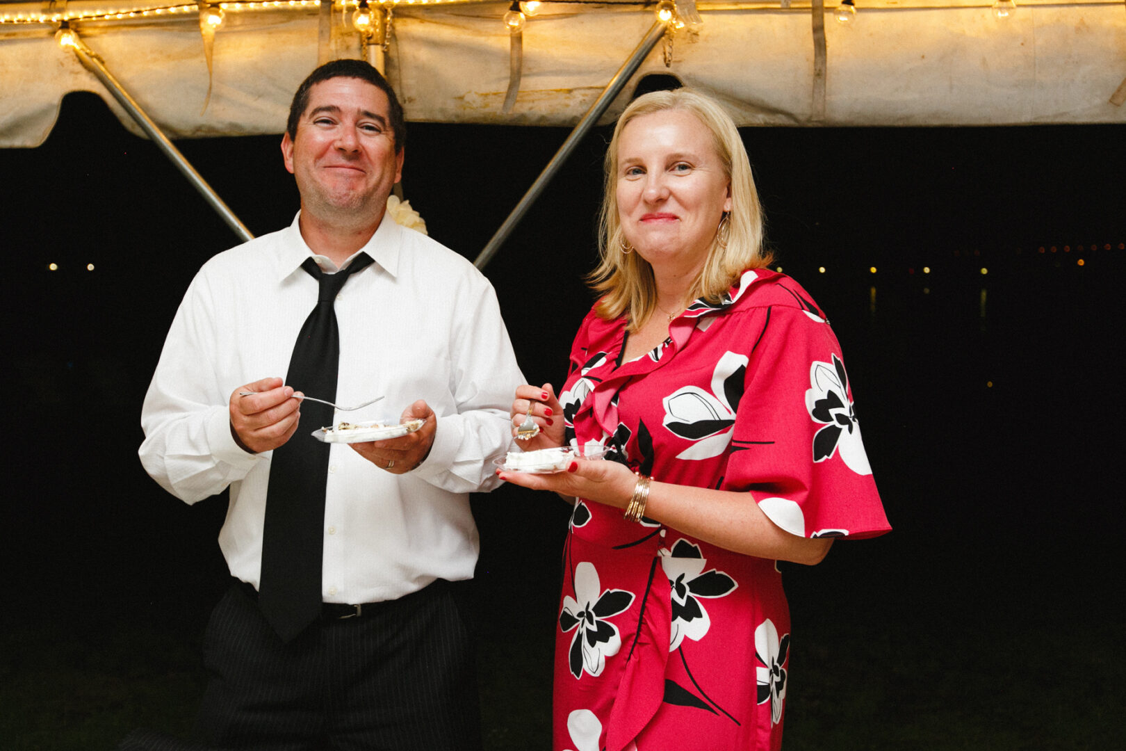 At a Corinthian Yacht Club of Philadelphia wedding, a man in a white shirt and tie and a woman in a floral dress beam as they hold dessert plates under a tent adorned with string lights.