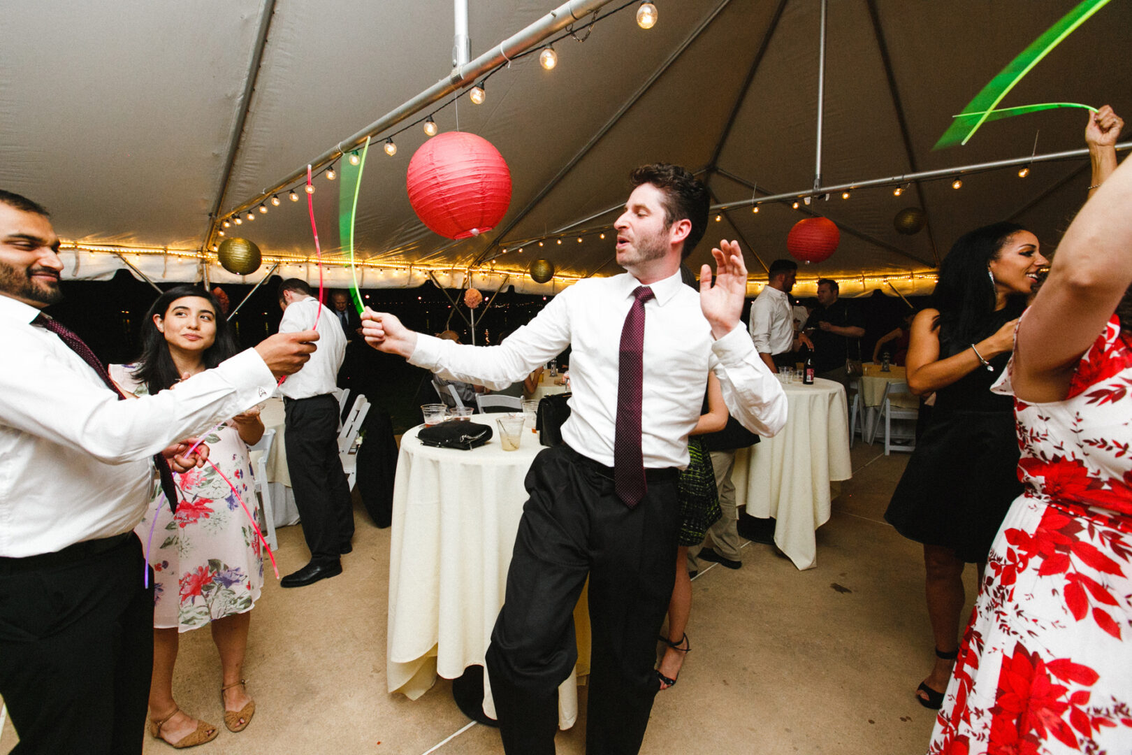 Amidst the lively atmosphere of a Corinthian Yacht Club of Philadelphia wedding, people danced under a tent, jubilantly waving colorful streamers. In the background, tables adorned with white tablecloths added an elegant touch to the festive scene.