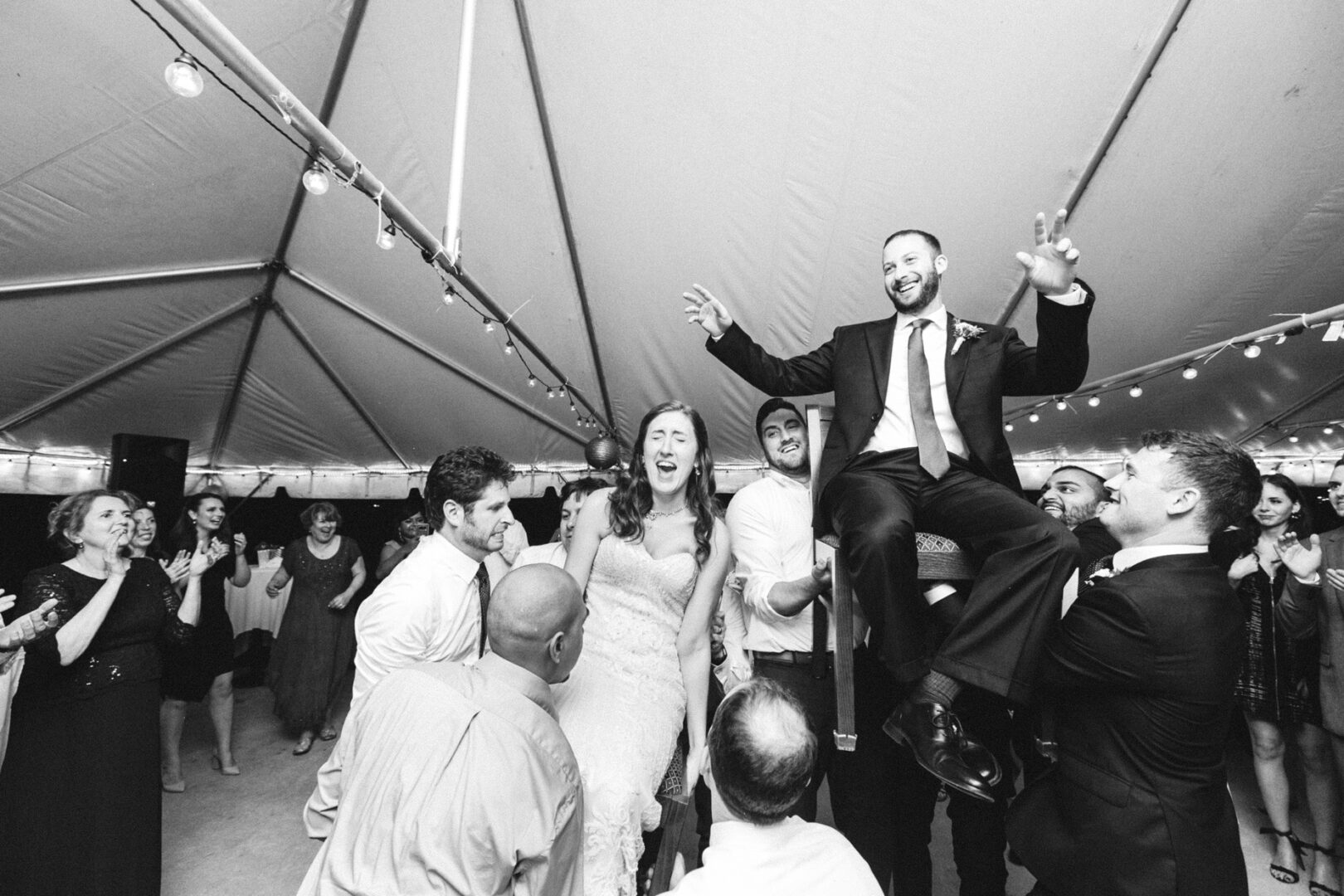 A black-and-white photo captures a Corinthian Yacht Club of Philadelphia wedding celebration under a tent, where a joyful couple is lifted on chairs by guests, amid dancing and laughter.