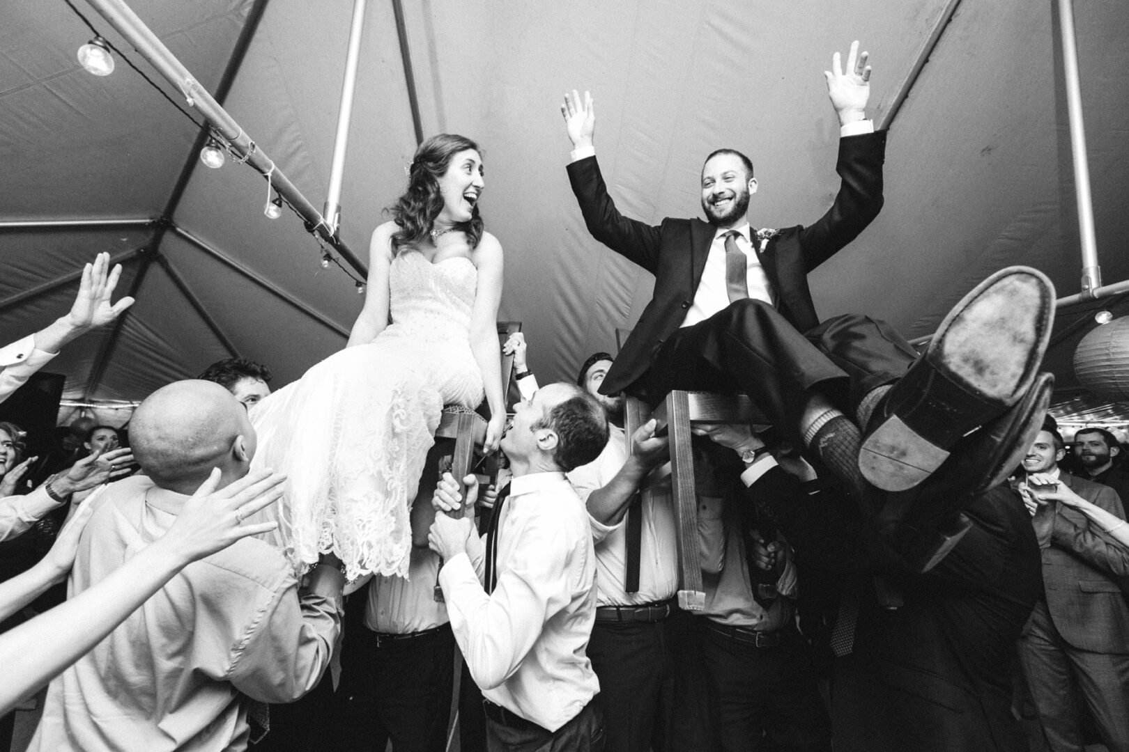 Amidst the joyous celebration under a tent at the Corinthian Yacht Club of Philadelphia, a bride and groom are lifted high on chairs, surrounded by cheering guests, making their special day unforgettable.