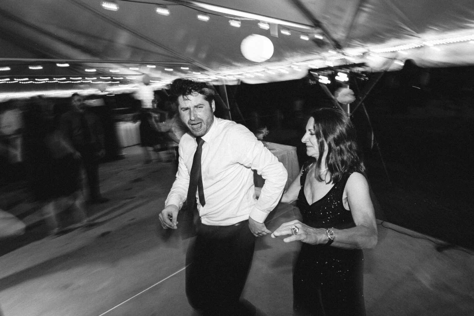 A man and a woman dance energetically under a tent adorned with string lights, capturing the vibrancy of a Corinthian Yacht Club of Philadelphia wedding. The black and white image adds a timeless elegance to the lively event.
