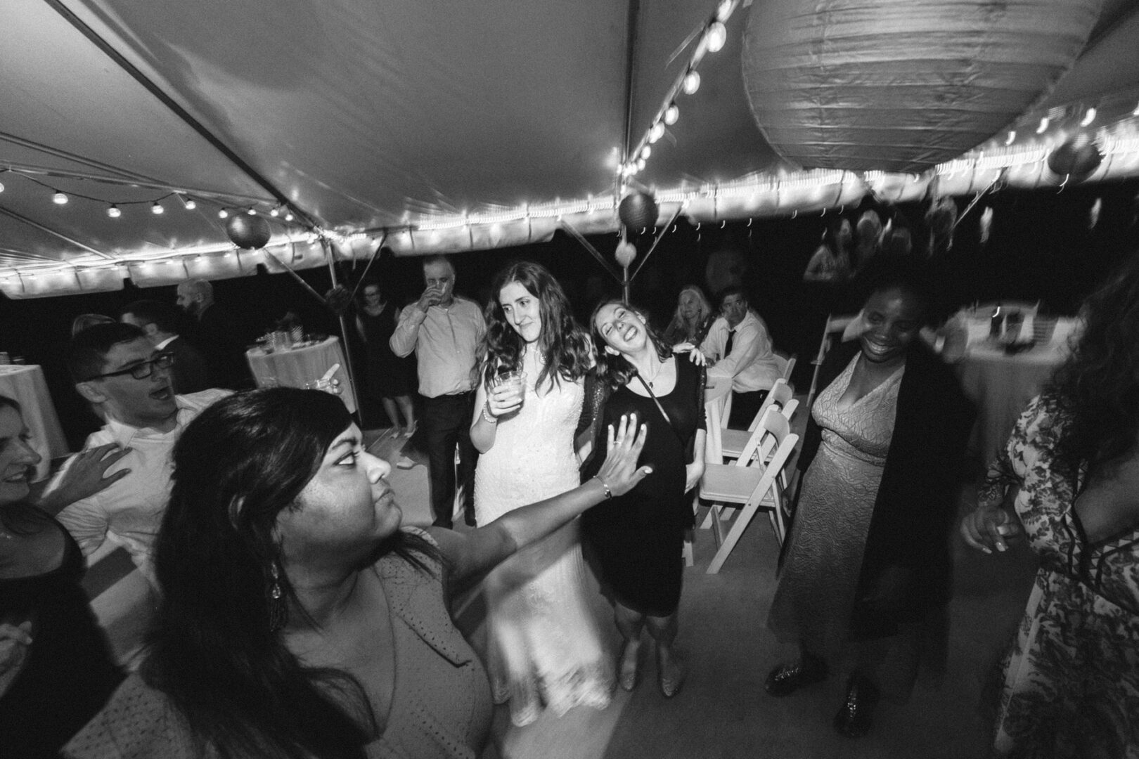 A group of people, including a person in a wedding dress, dance and socialize under twinkling string lights at the Corinthian Yacht Club of Philadelphia wedding, creating an enchanting tented event.