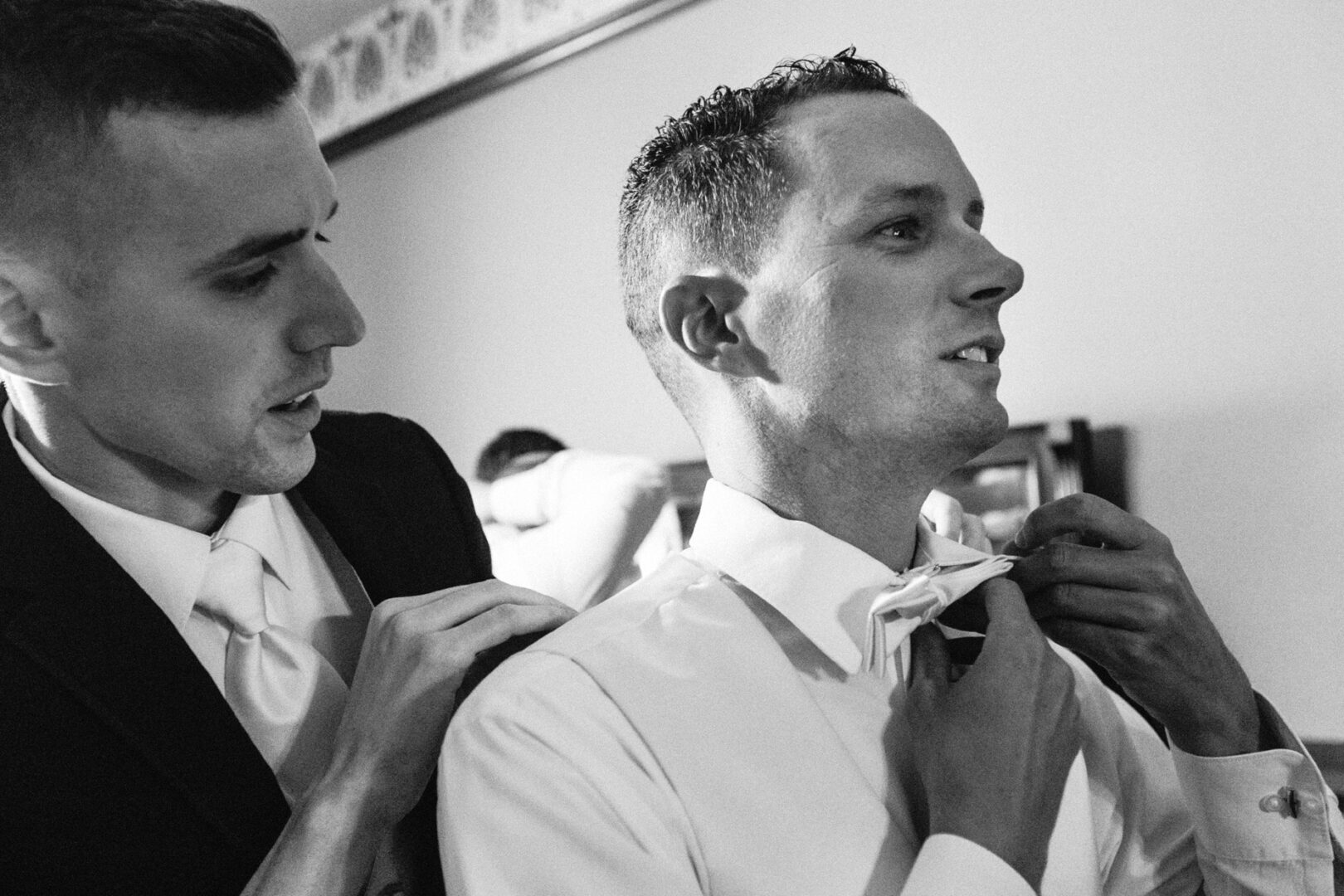 In a room with patterned wallpaper, a man assists another in adjusting his bow tie, both impeccably dressed in suits, perhaps preparing for the elegance of a Knowlton Mansion wedding.