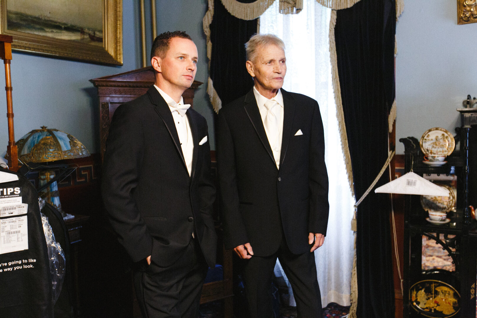 Two men in formal black suits stand in a vintage room with dark curtains and antique furnishings, reminiscent of a Knowlton Mansion wedding.