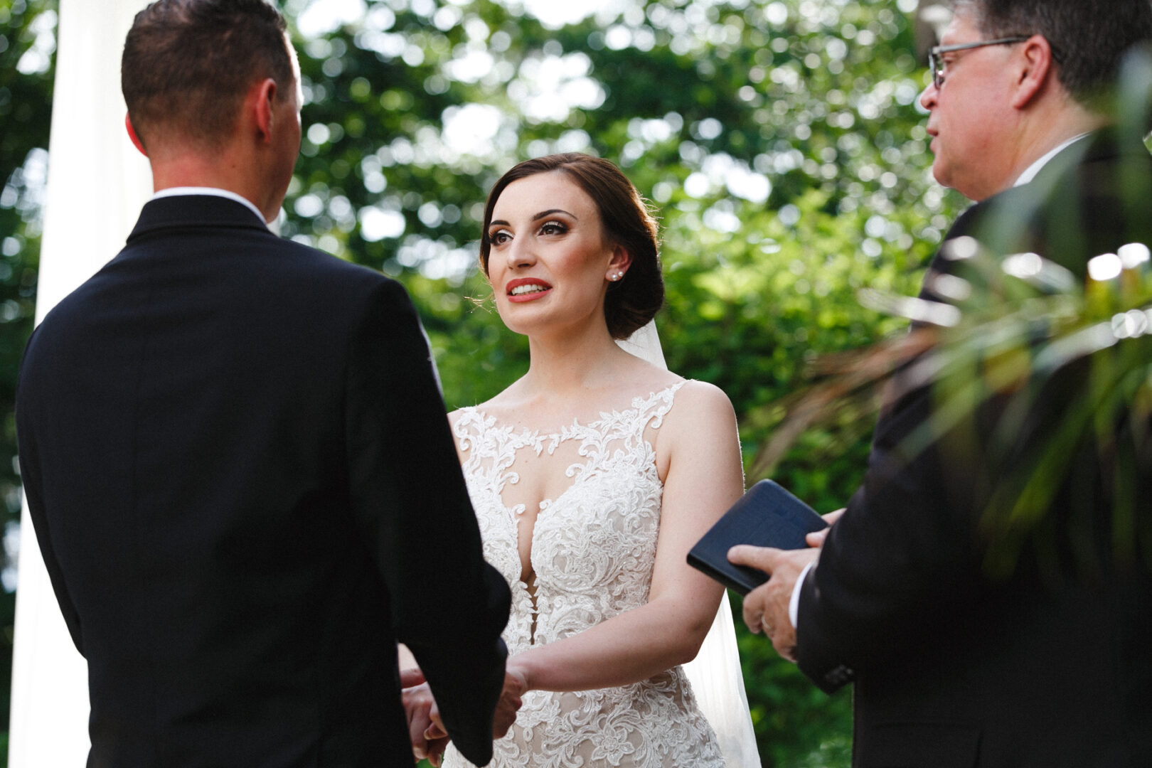 At a picturesque Knowlton Mansion Wedding, a woman in a white dress holds hands with a man in a suit as an officiant stands nearby, amidst the charming outdoor setting.