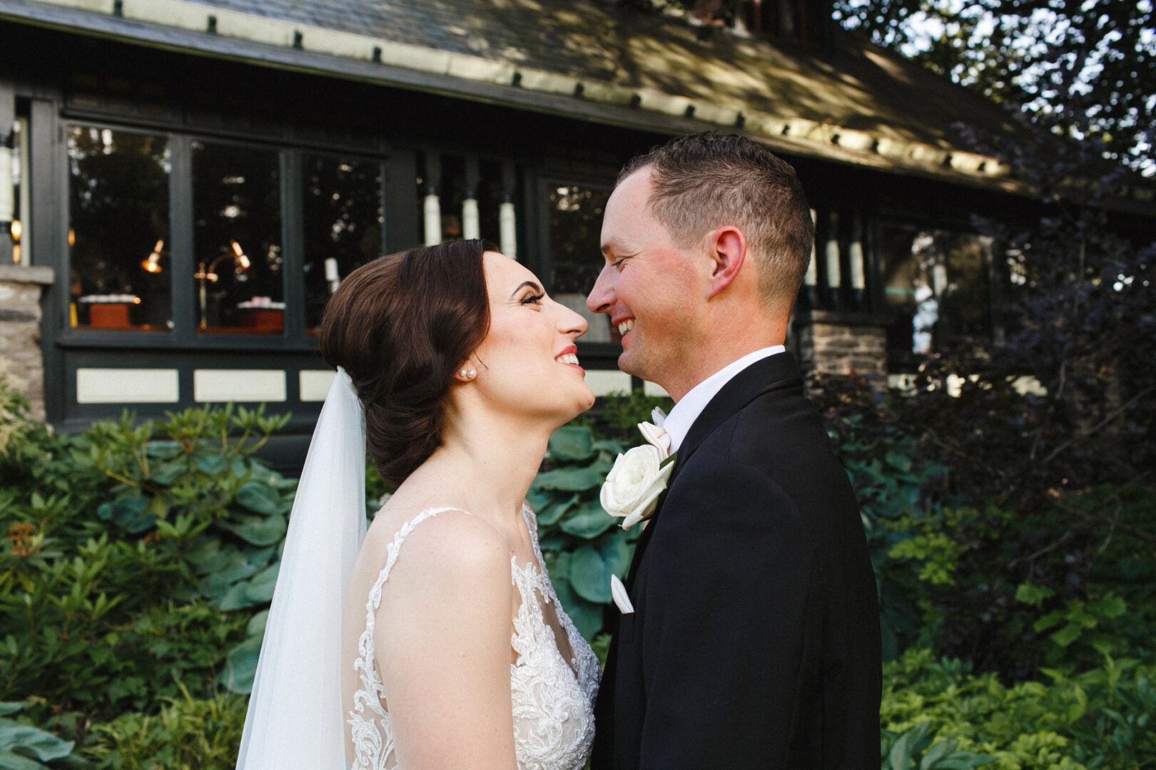 A bride and groom in wedding attire smile at each other, captured beautifully amidst the greenery of Knowlton Mansion Wedding grounds, with a charming building in the background.
