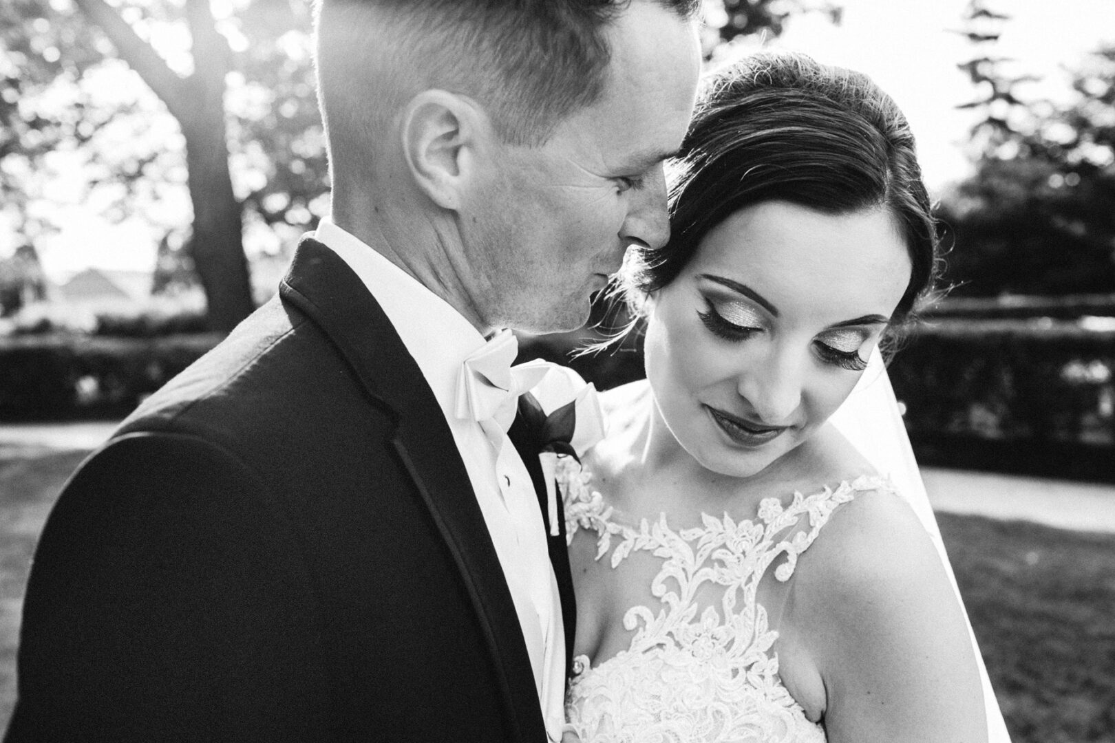 A bride and groom pose intimately outdoors at their Knowlton Mansion wedding. The bride wears a lace dress, and the groom is in a suit with a bow tie. Sunlight filters through the trees in the background, casting a romantic glow over their special day.