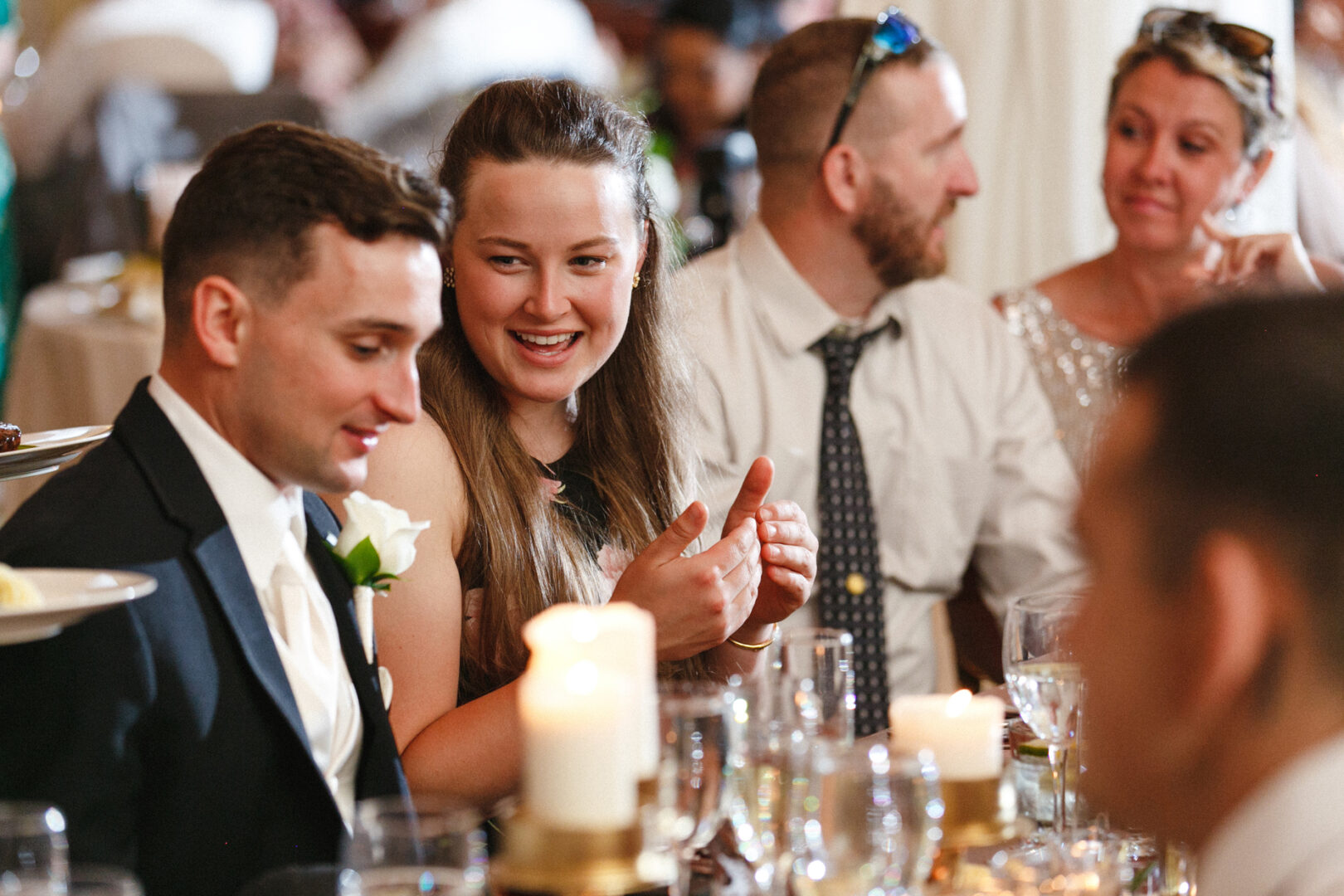 At a Knowlton Mansion wedding, a group of people dressed formally sits at a dining table with lit candles and glasses. One person is clapping, while others are engaged in conversation, adding to the festive atmosphere.