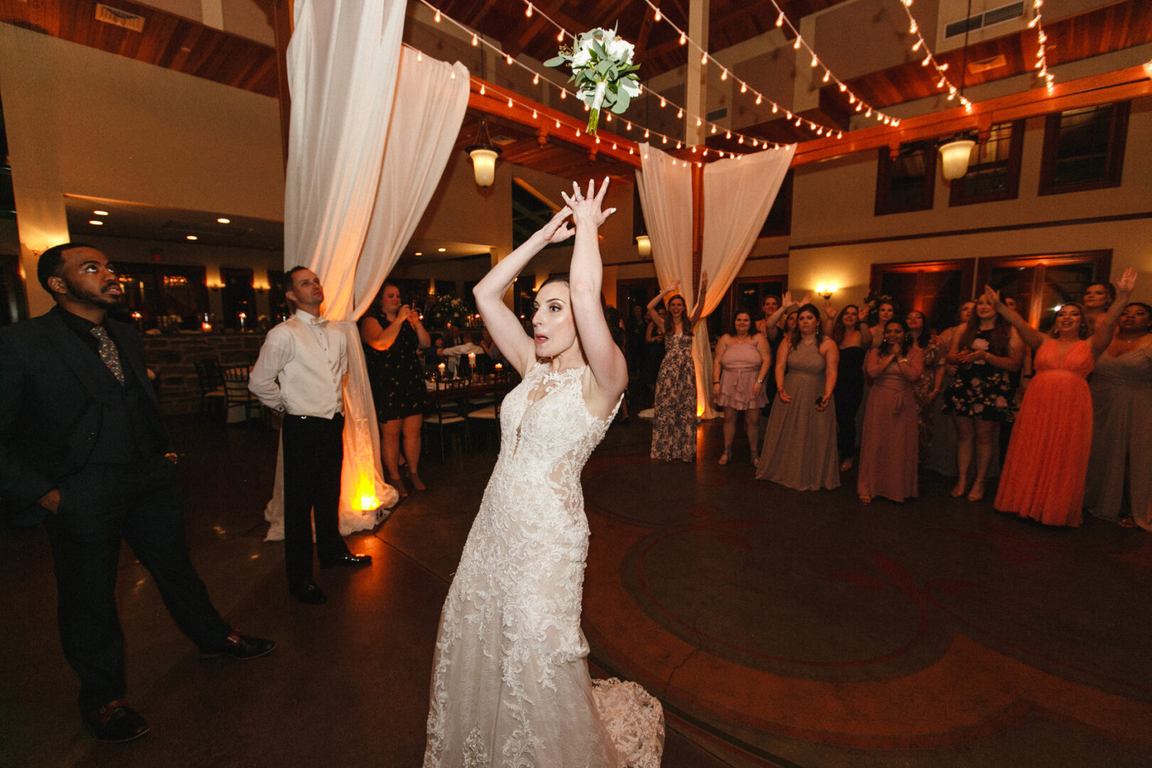 At a Knowlton Mansion Wedding, a bride in a white dress tosses her bouquet over her head towards a group of eager women. Guests watch the scene unfold beneath elegant draped fabric and twinkling string lights.