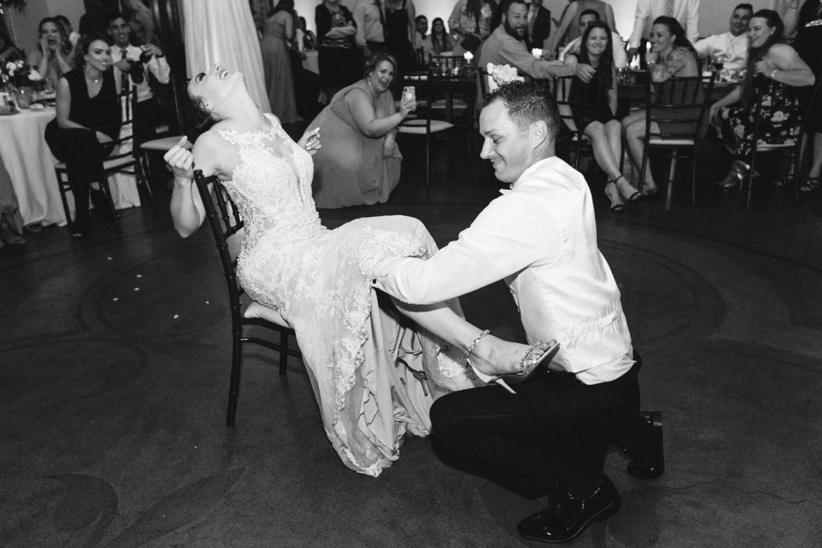 At a charming Knowlton Mansion wedding, the groom kneels to remove a garter from the bride's leg during the reception. Guests seated at elegantly decorated tables watch and react in delight in the background.