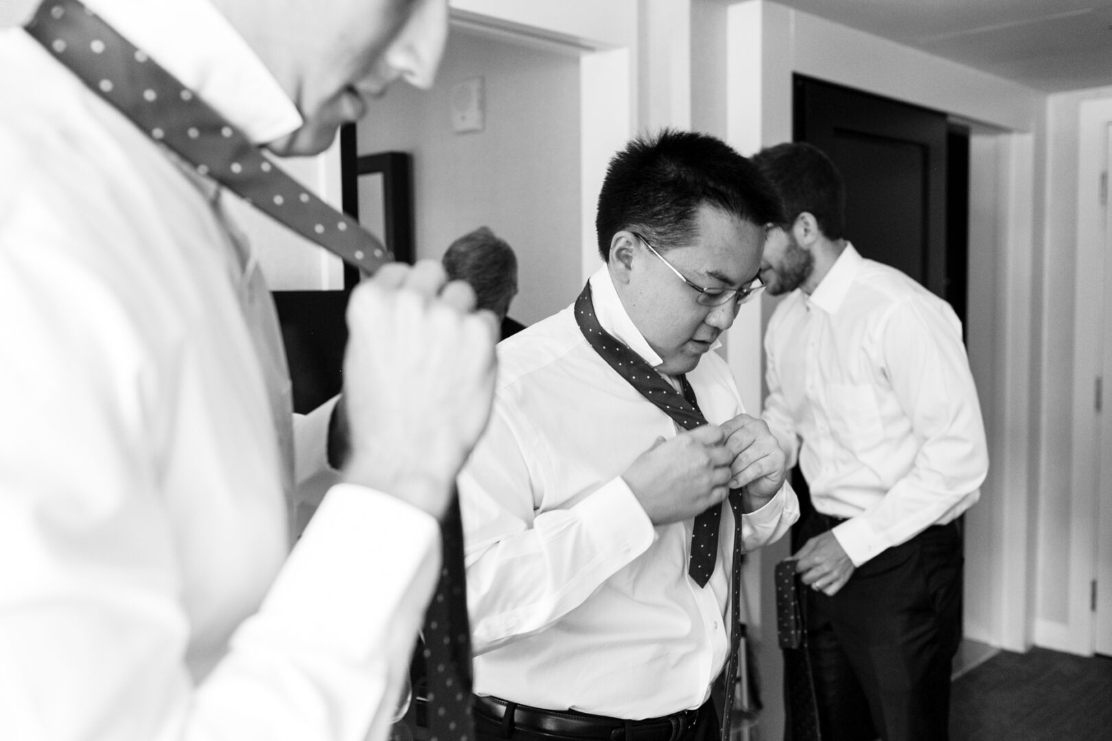 Three men in white shirts are tying neckties in a room, preparing for a Colonial Dames wedding. Two are visible in the foreground, concentrating on their ties, while one stands in the background adjusting his tie with precision.