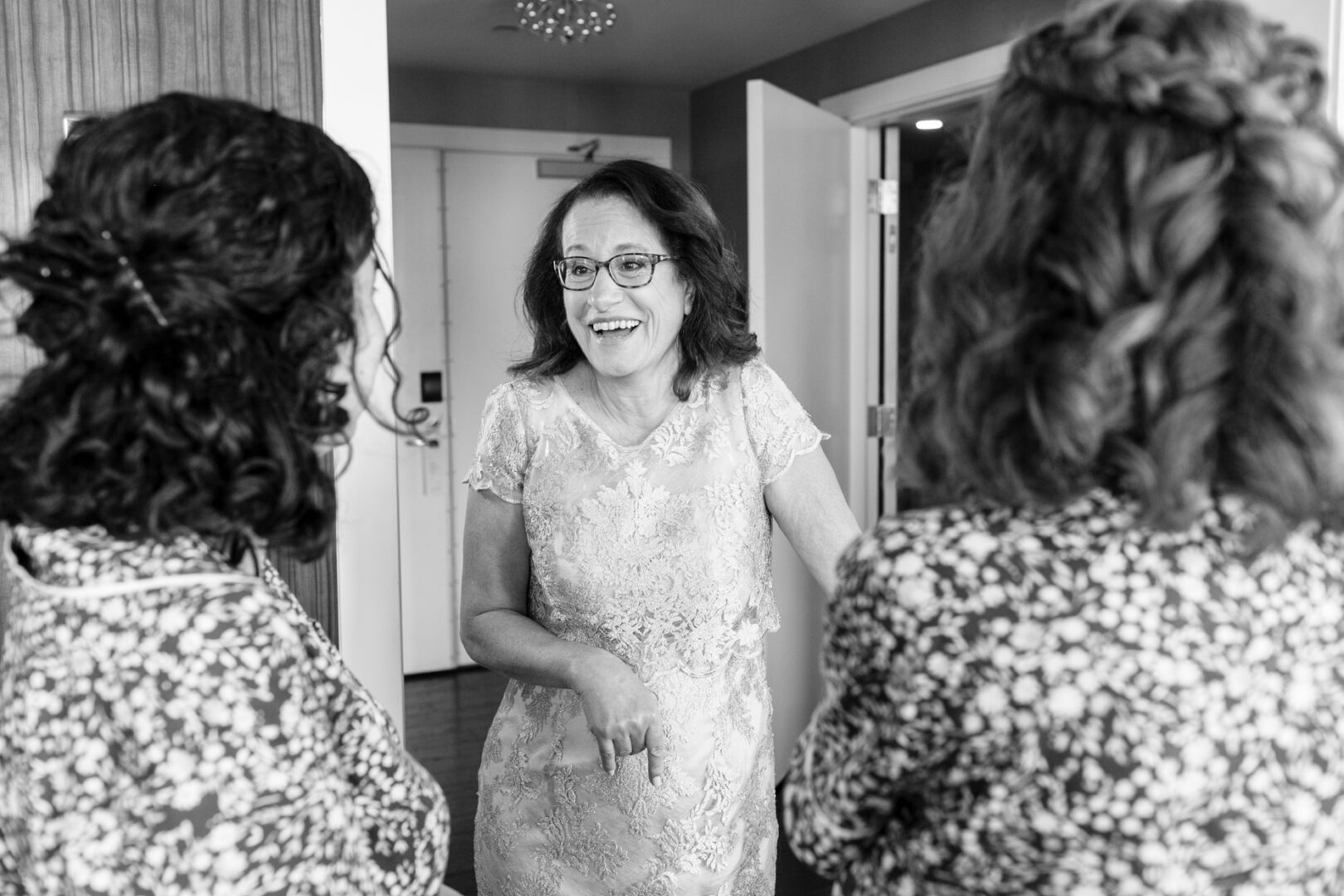 In a room reminiscent of a Colonial Dames gathering, a woman in glasses and a lace dress smiles while engaging with two people adorned in floral dresses, as if delighted guests at an intimate wedding celebration.