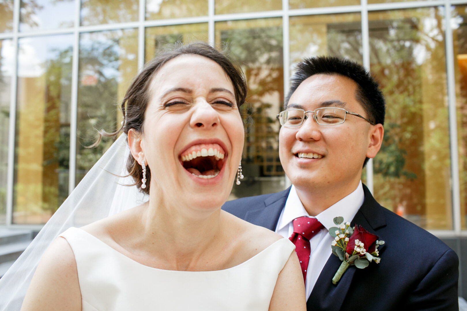 Bride laughing while standing next to groom in a suit with a red tie and boutonniere, outside the elegant setting of a Colonial Dames wedding venue, surrounded by glass windows.