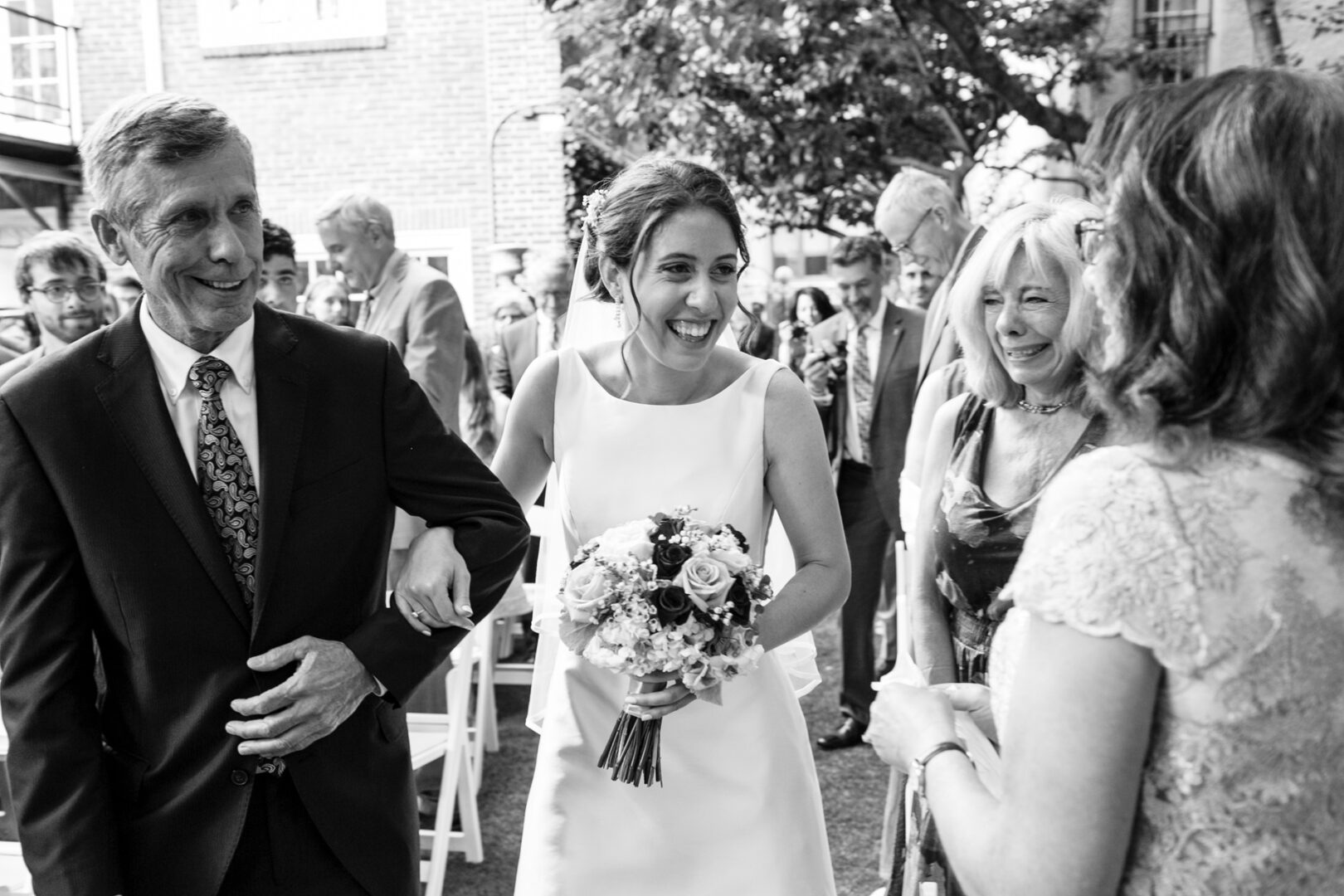 At a charming Colonial Dames wedding, a bride in a white dress holds a bouquet, smiling and linking arms with a man in a suit. They are surrounded by delighted guests outdoors.