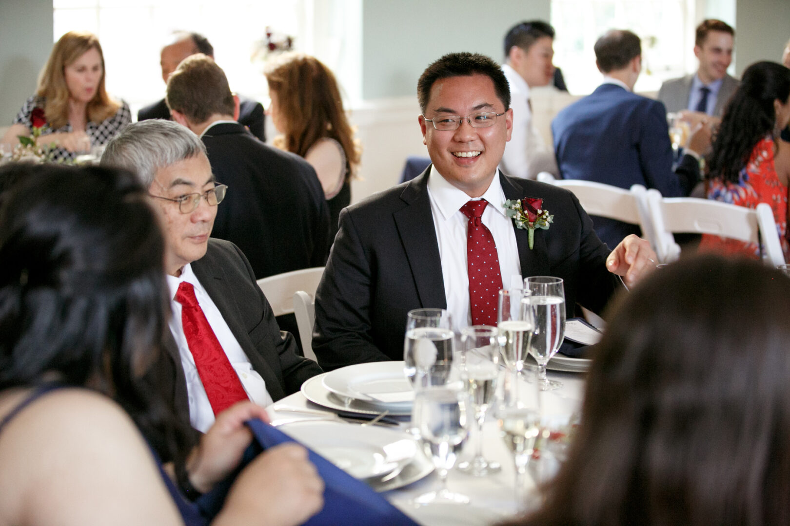 A smiling man in a suit sits at a formal table with guests, evoking the elegance of a Colonial Dames wedding. Plates and glasses are meticulously arranged on the table.