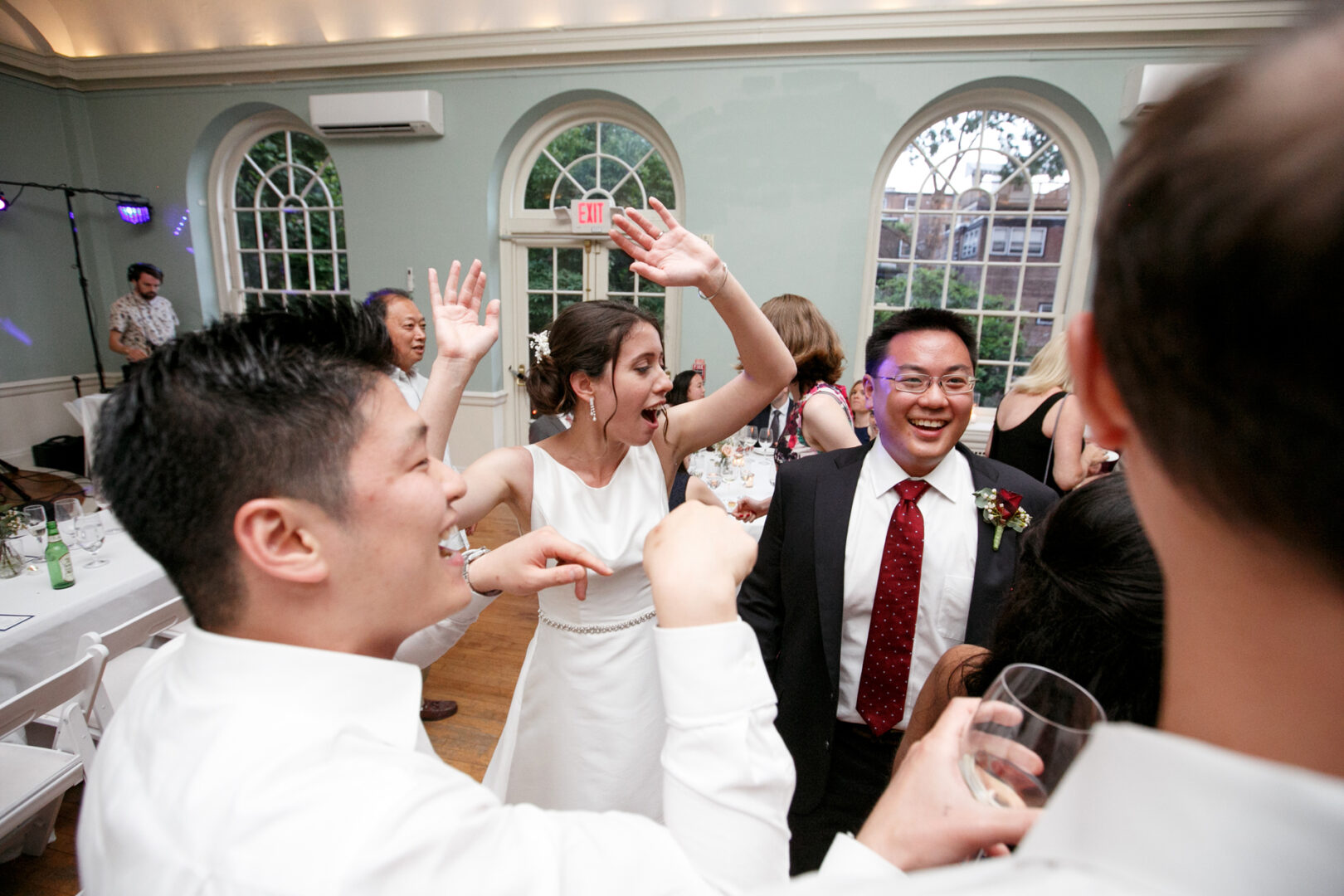 Wedding guests dance and celebrate in a Colonial Dames hall with arched windows. A woman in a white dress raises her arms, while a man in a suit smiles.