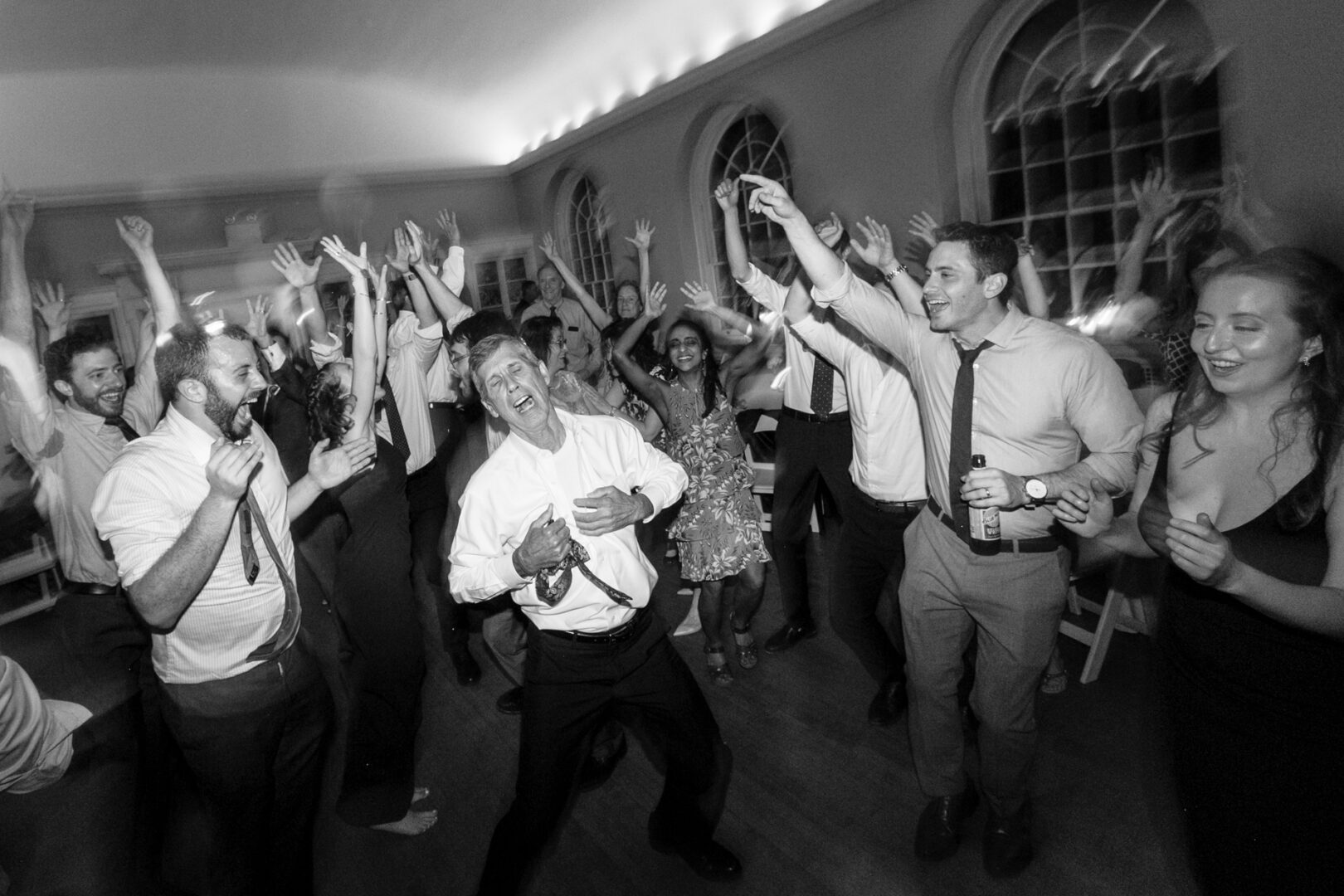 A lively group of people, reminiscent of Colonial Dames elegance, dancing and cheering in a crowded room, with several raising their hands in the air, captured beautifully in black and white.