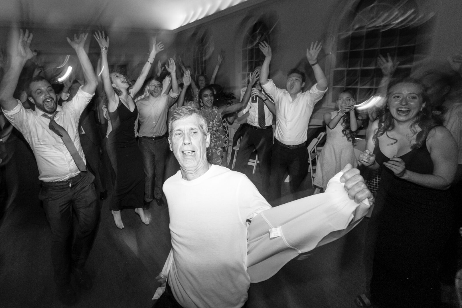 Amidst a Colonial Dames wedding, a crowd of people dance energetically with their hands raised and joyful expressions in a room with arched windows. A man in a white shirt stands prominently in the foreground, embodying the spirit of celebration.