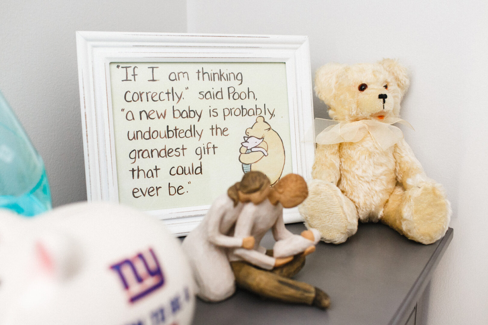 Framed Pooh quote about a new baby, surrounded by newborn photos, with a teddy bear and porcelain figurine on a shelf.