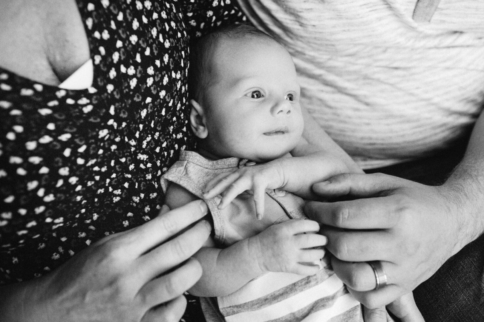 A newborn lies peacefully on an adult's lap, surrounded by caring hands. The baby gazes up, clad in a striped outfit, while the adults don patterned and striped shirts. This black and white image captures the timeless essence of newborn photos.