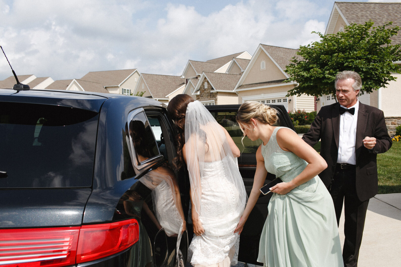 A bride in a white dress and veil enters a black car assisted by a bridesmaid, capturing the serene charm of a High Point wedding. Nearby, an older man in a black tuxedo stands in the residential neighborhood, adding to the moment's elegance.