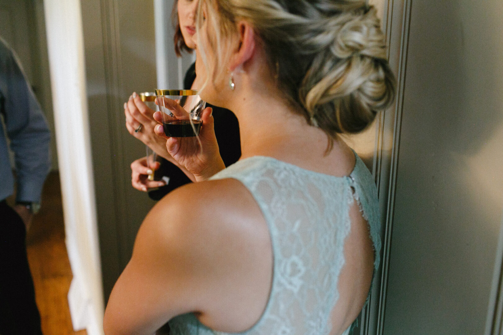 Woman in lace dress holding a drink, engaged in conversation with a partially visible person with reddish hair near a wall at the High Point Wedding.