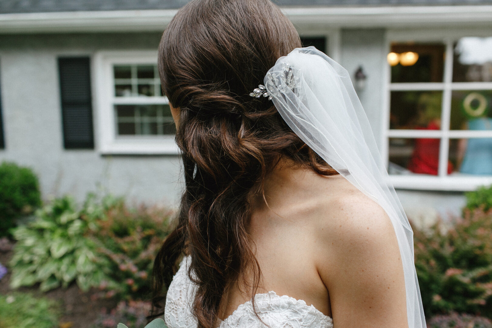 The bride, her long hair cascading down her back, stands outdoors at High Point Wedding. She's in a strapless white dress with a delicate veil, facing away from the camera.