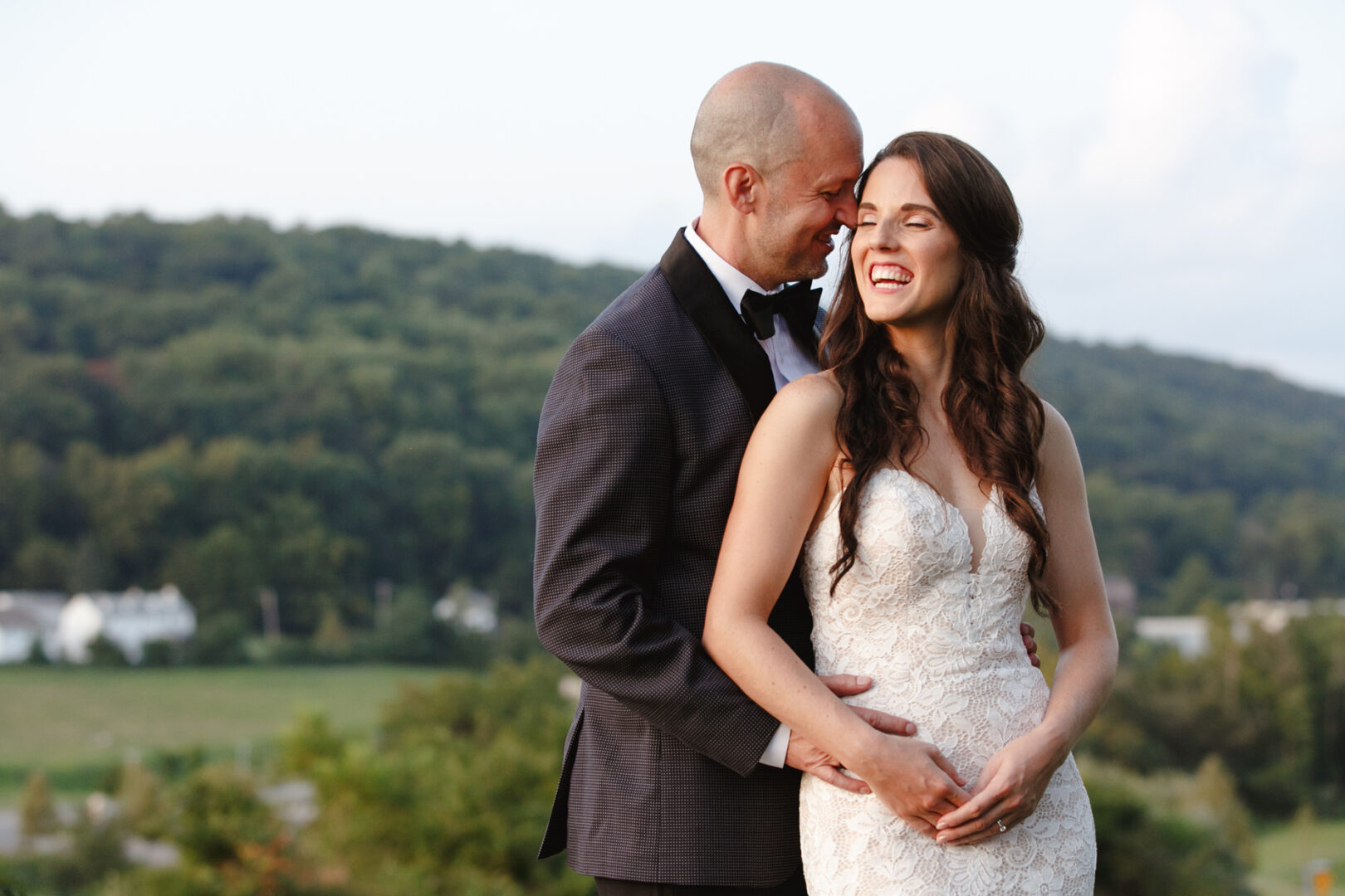A couple in wedding attire stands amidst the scenic beauty of High Point, the man embracing the woman from behind. She is smiling with eyes closed, surrounded by the gentle embrace of a hilly landscape in the background.