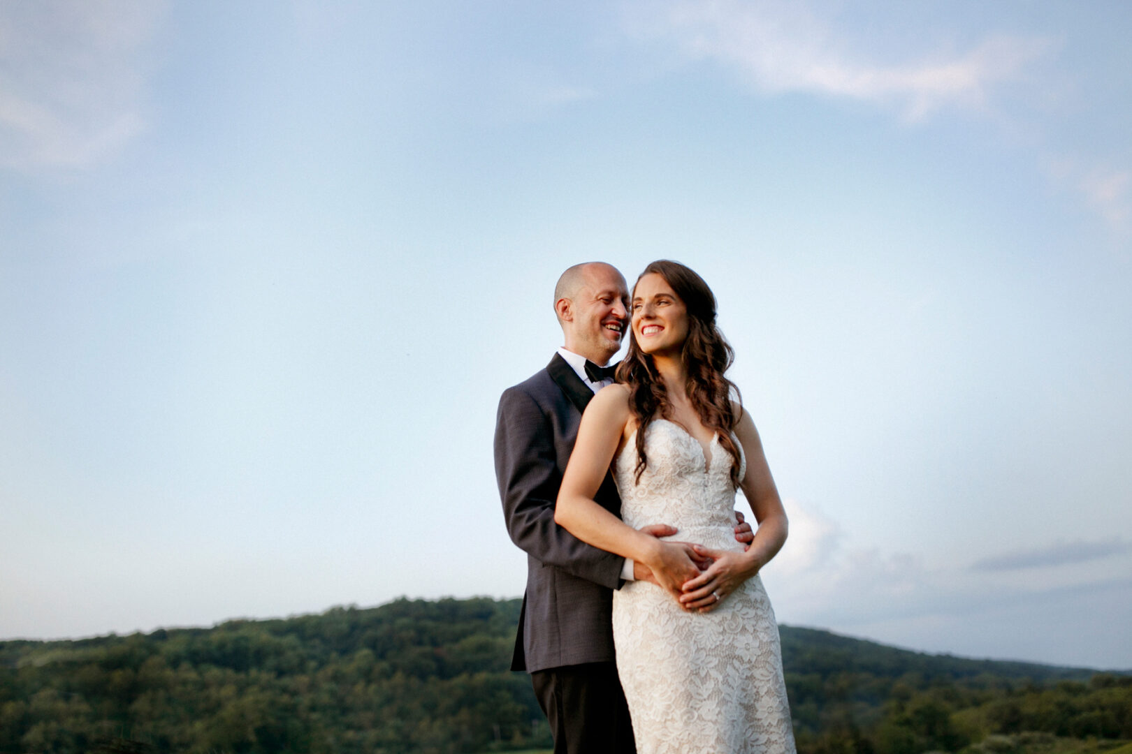 A couple, standing outdoors at a high point, poses for a photo against a scenic backdrop with hills and a clear sky. The woman is in a white dress, and the man is wearing a suit, capturing their wedding's enchanting essence.