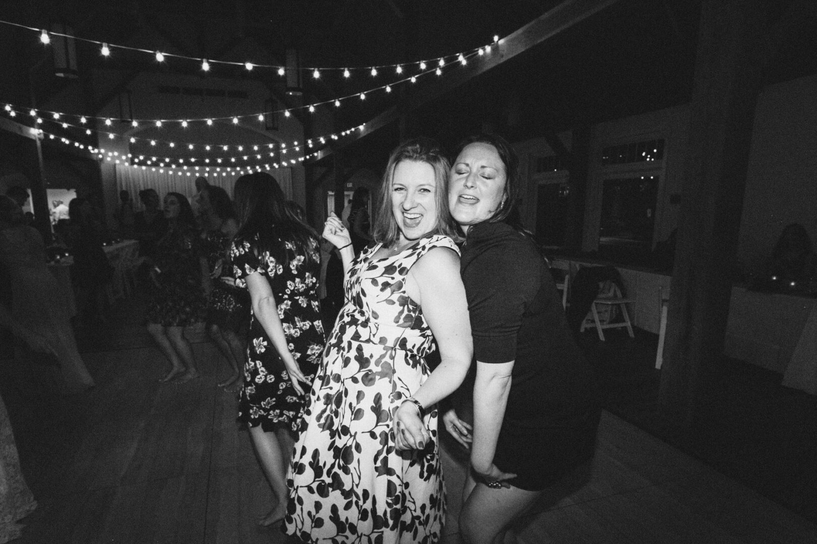 Two women dance joyfully under string lights at a High Point Wedding, surrounded by other attendees. The scene captures a lively and festive atmosphere.