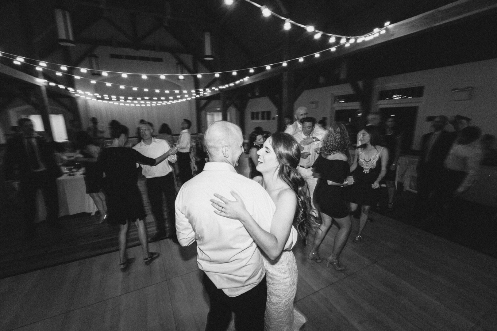 A couple sways gracefully in a dimly lit room with string lights casting a warm glow, reminiscent of a High Point Wedding. In the background, guests mingle and dance, adding to the enchanting atmosphere.