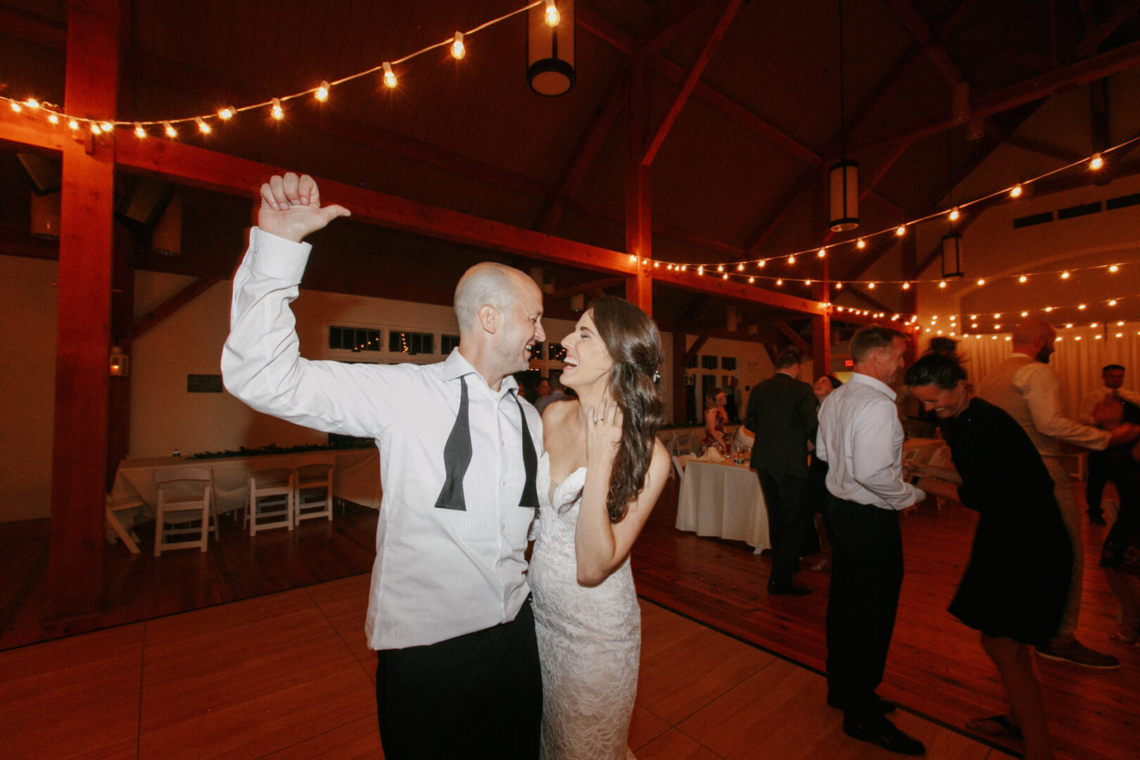Bride and groom dancing joyfully under string lights in a rustic wooden hall at their charming High Point Wedding, with guests celebrating in the background.