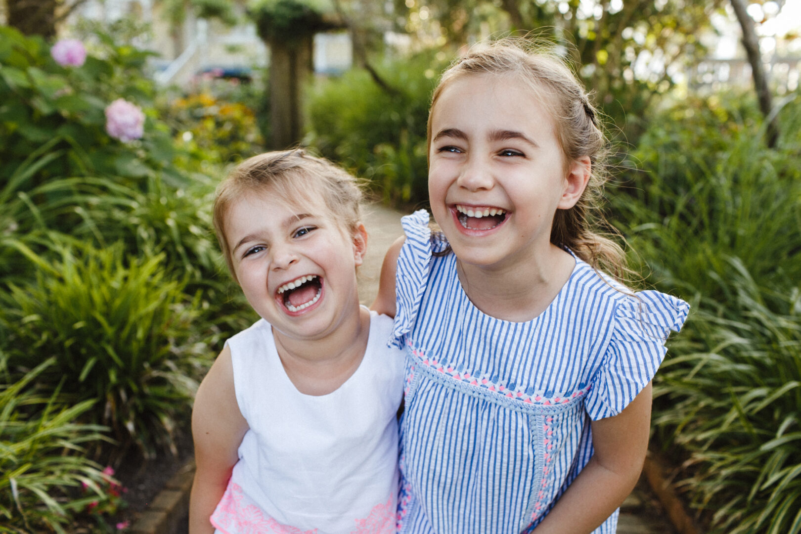Two young girls smiling broadly while standing close together in a garden with green foliage in the background, capturing a moment reminiscent of joyful Jersey Shore family photos.