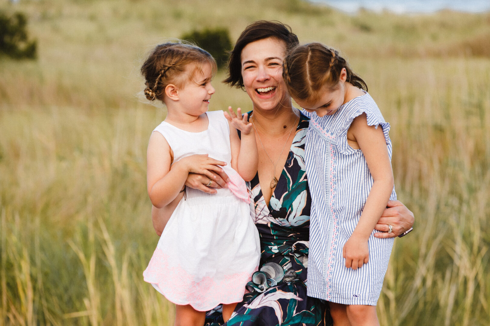 A woman in a floral dress holds two young girls in white dresses, creating a picture-perfect moment reminiscent of Jersey Shore family photos, as they stand together in a grassy field.