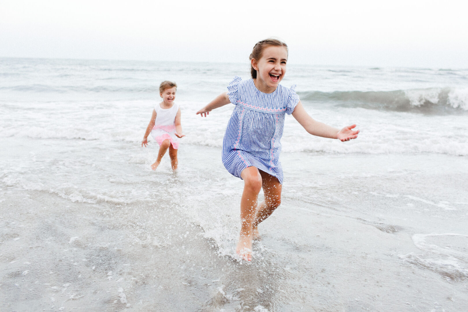 Two young girls joyfully run and splash in the shallow ocean waves at the Jersey Shore on a cloudy day, their summer dresses twirling with each playful step.