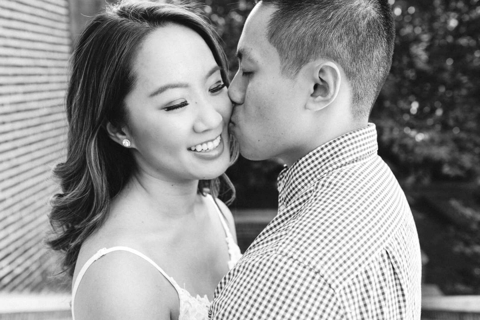 A man softly kisses the cheek of a smiling woman, their happiness captured in a black and white moment. Standing close outdoors on a sunny day, their affection hints at a University of Pennsylvania engagement.