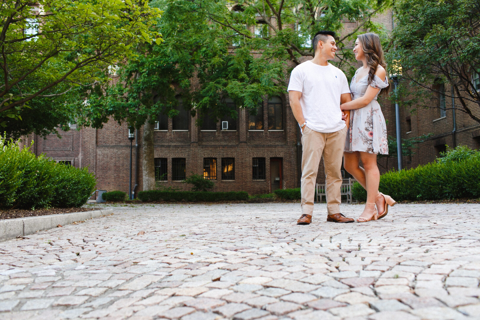 A couple stands on a cobblestone path in front of a brick building, surrounded by greenery at the University of Pennsylvania. They face each other, with the woman gently holding the man's arm, capturing a sweet engagement moment.