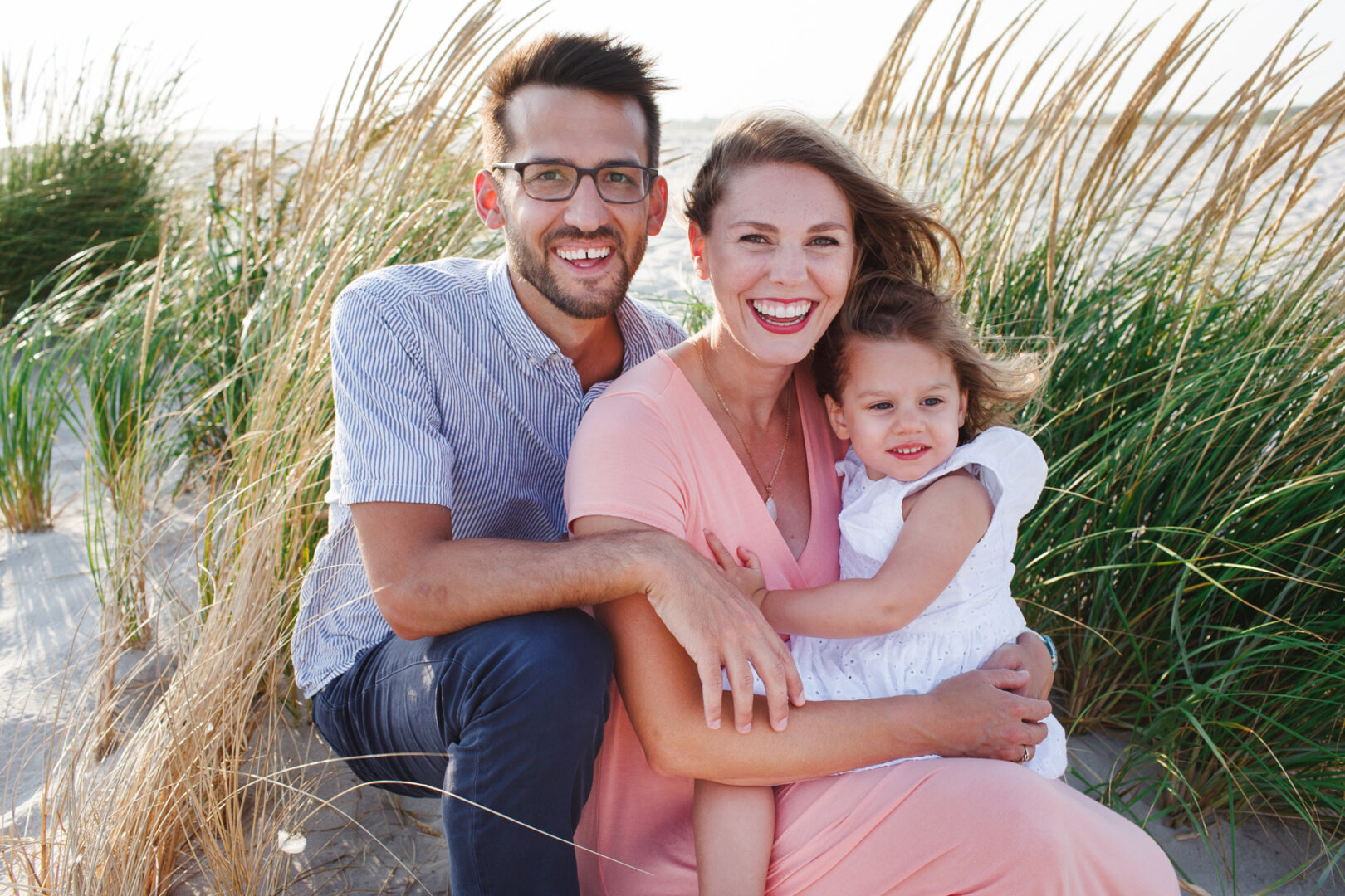 A man, woman, and child sit together on the sand at Cape May, surrounded by grass. The woman cradles the child as they all beam at the camera, capturing a perfect moment in their family photo.