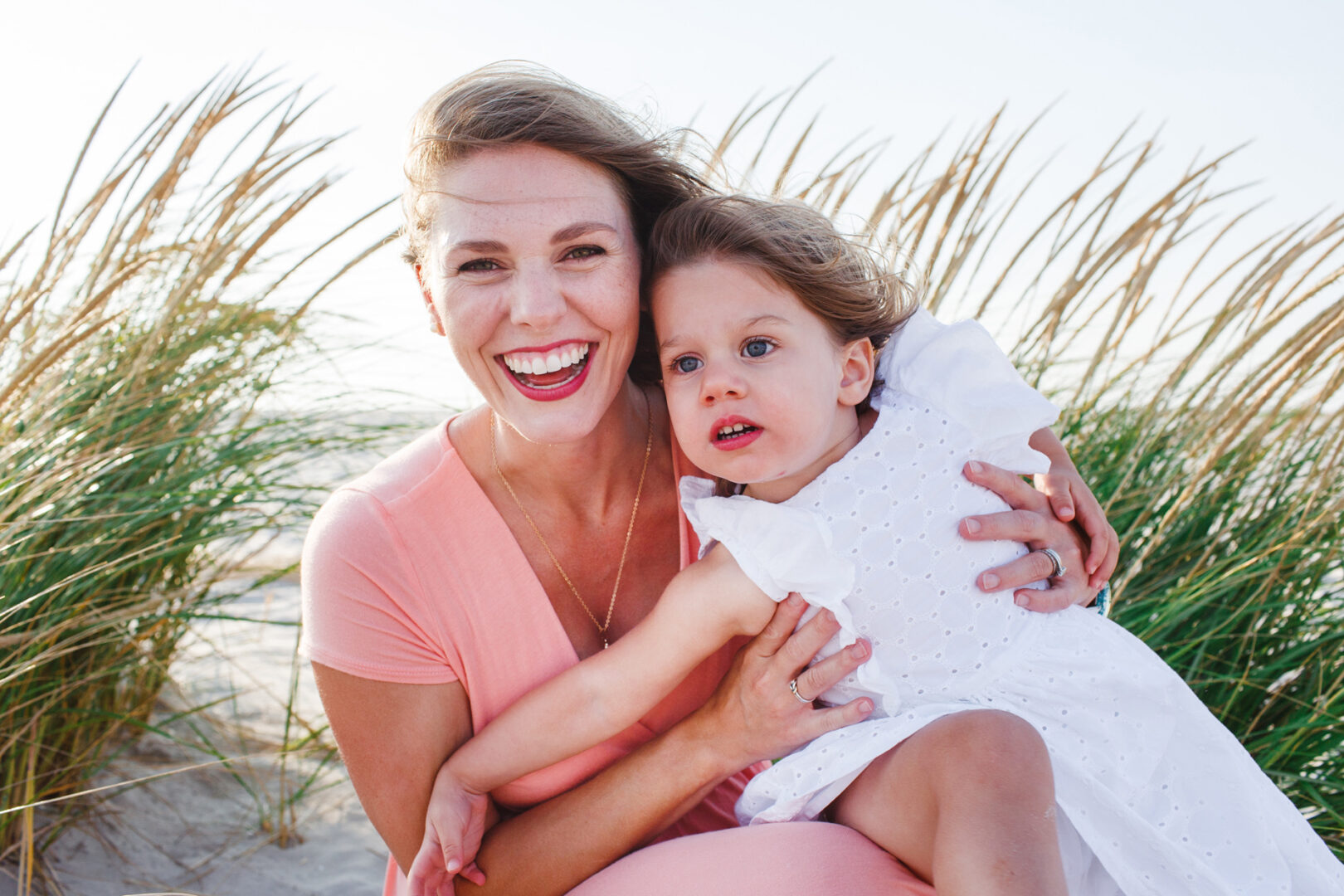 A woman and a child sit on a beach in Cape May, surrounded by tall grass. The woman is smiling broadly, while the child looks content, capturing the essence of cherished family photos.