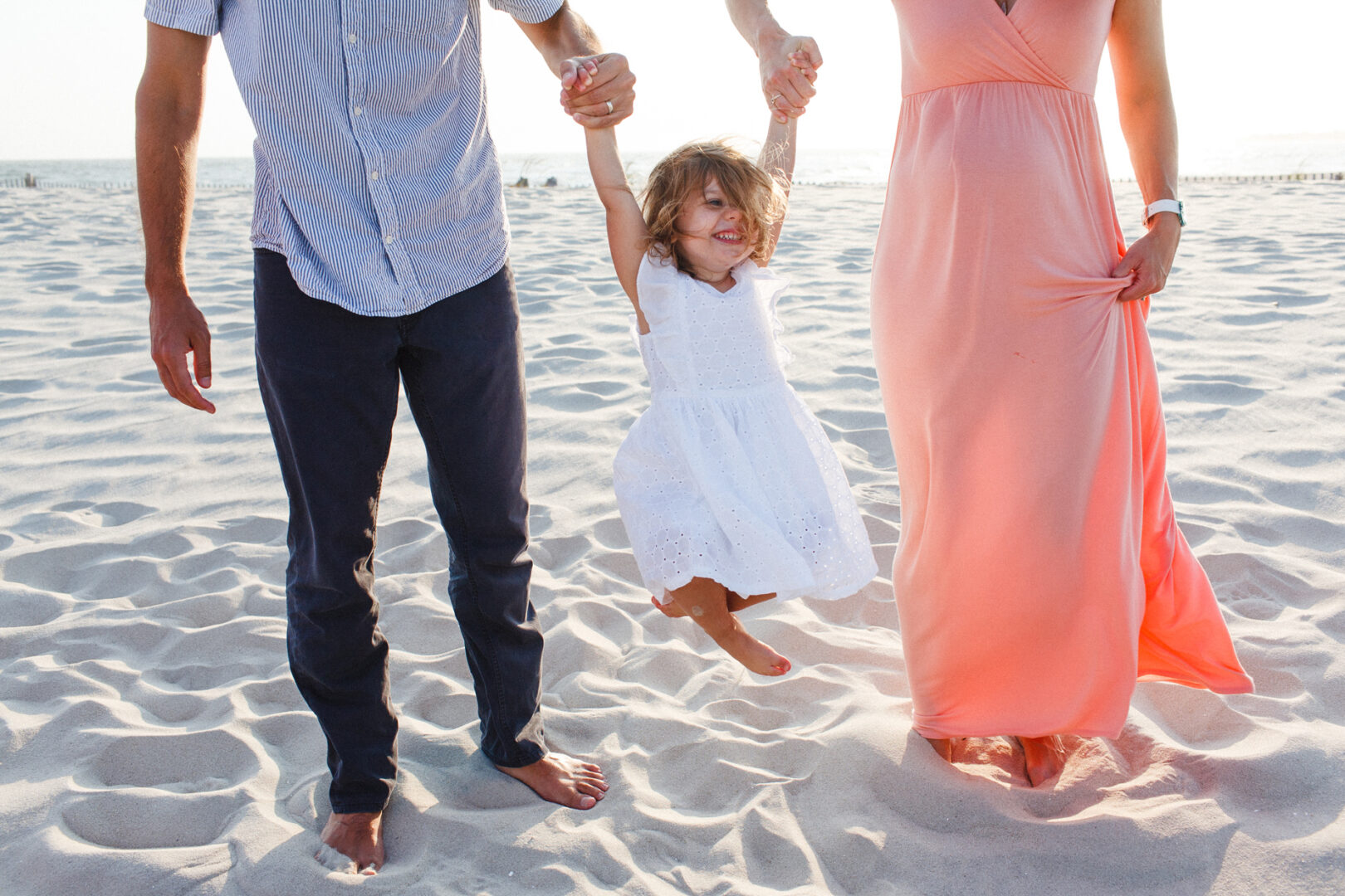 At Cape May, a child in a white dress is playfully lifted by two adults on the sandy beach, capturing a perfect family photo moment.