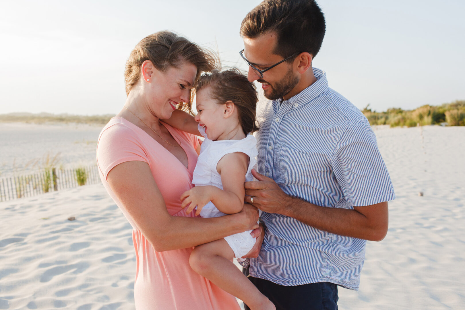 A family of three smiles and embraces on a sandy Cape May beach. The woman wears a pink dress, the man a striped shirt, and the child a white dress. Dunes and clear skies create the perfect backdrop for these cherished family photos.